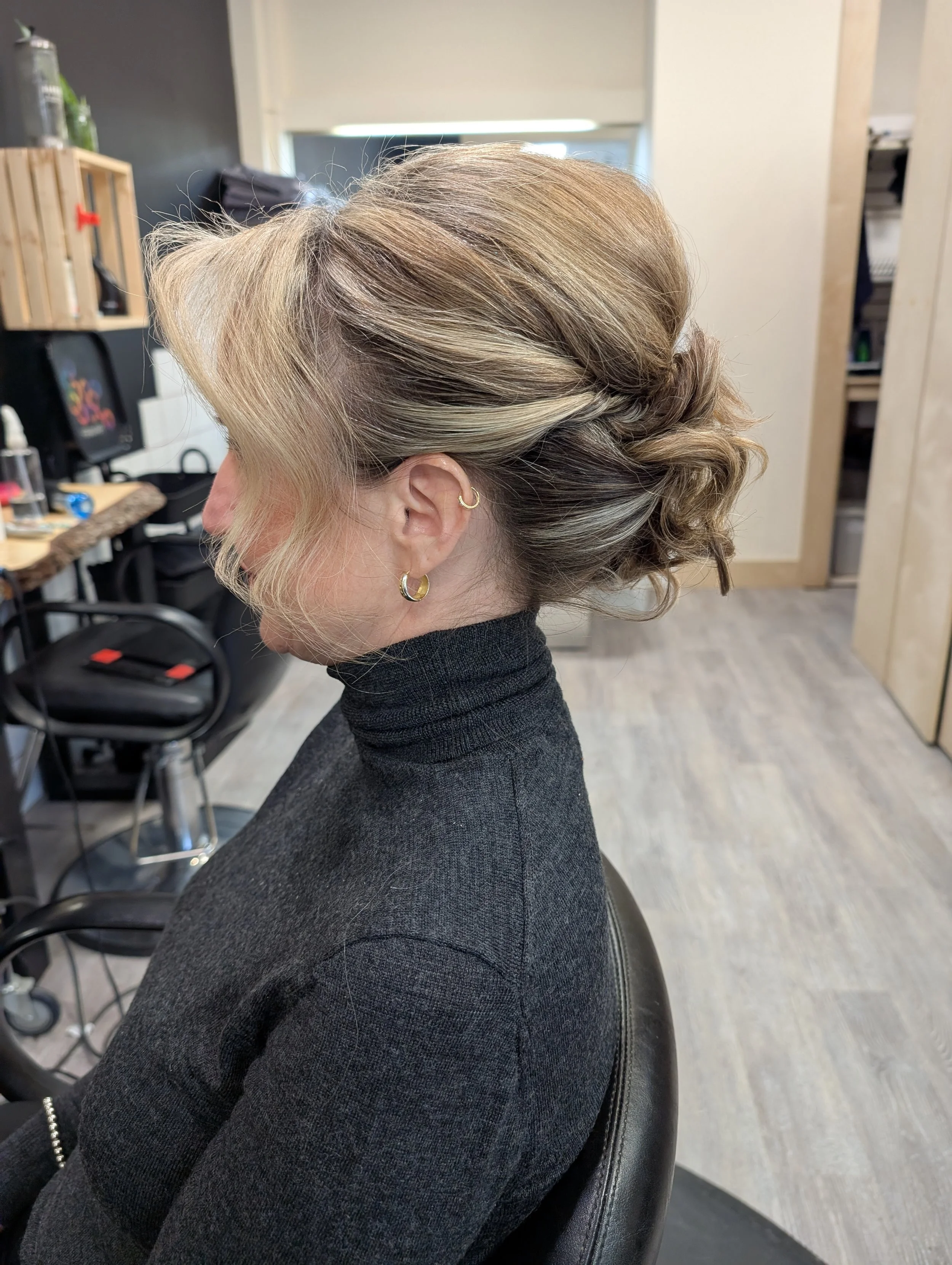 A woman with blonde, styled hair in an updo, wearing gold hoop earrings and a black turtleneck, sitting in a hair salon.