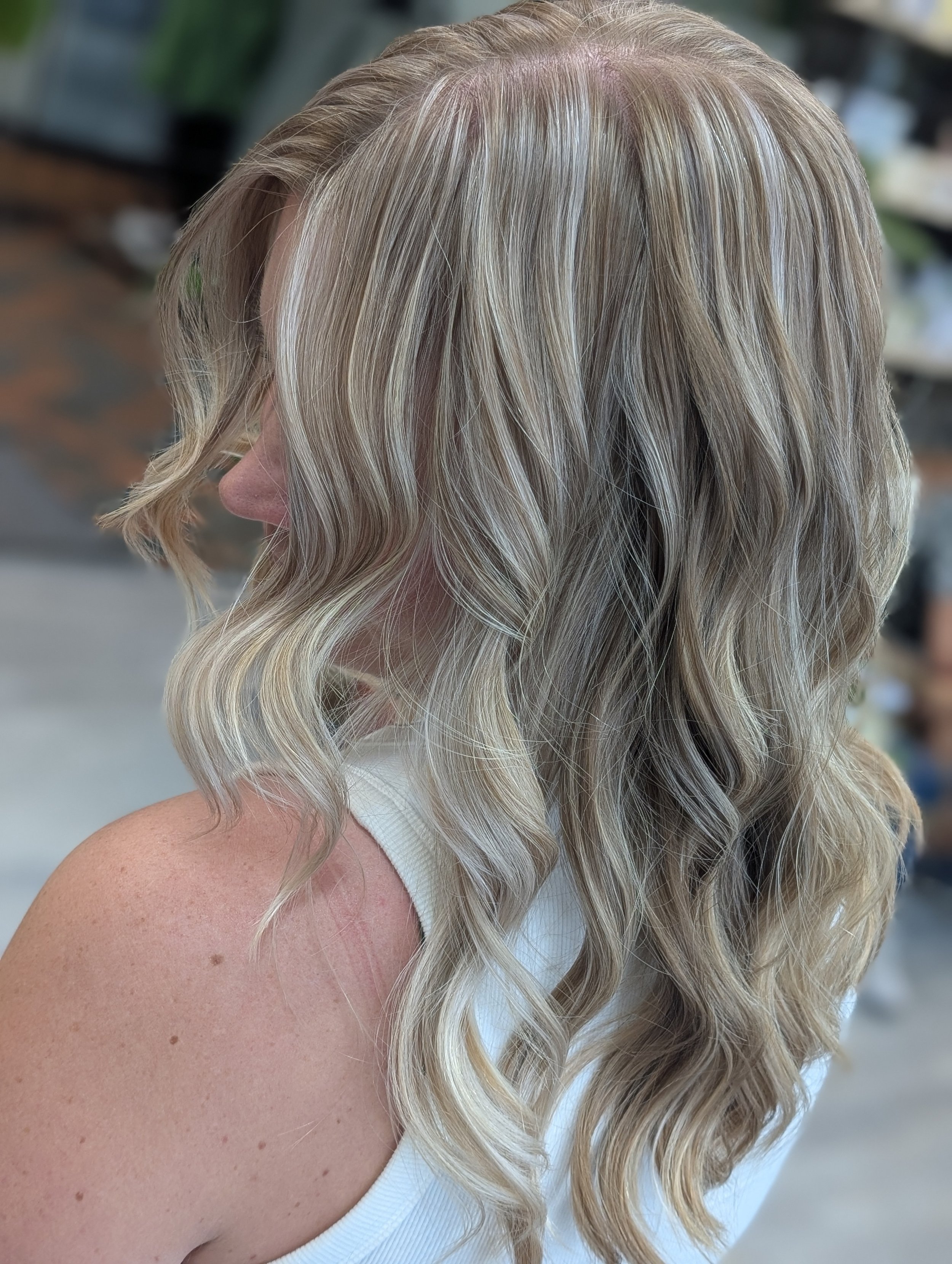 Close-up of a woman with long, blonde, wavy hair styled in loose curls, wearing a white top, with a blurred background.