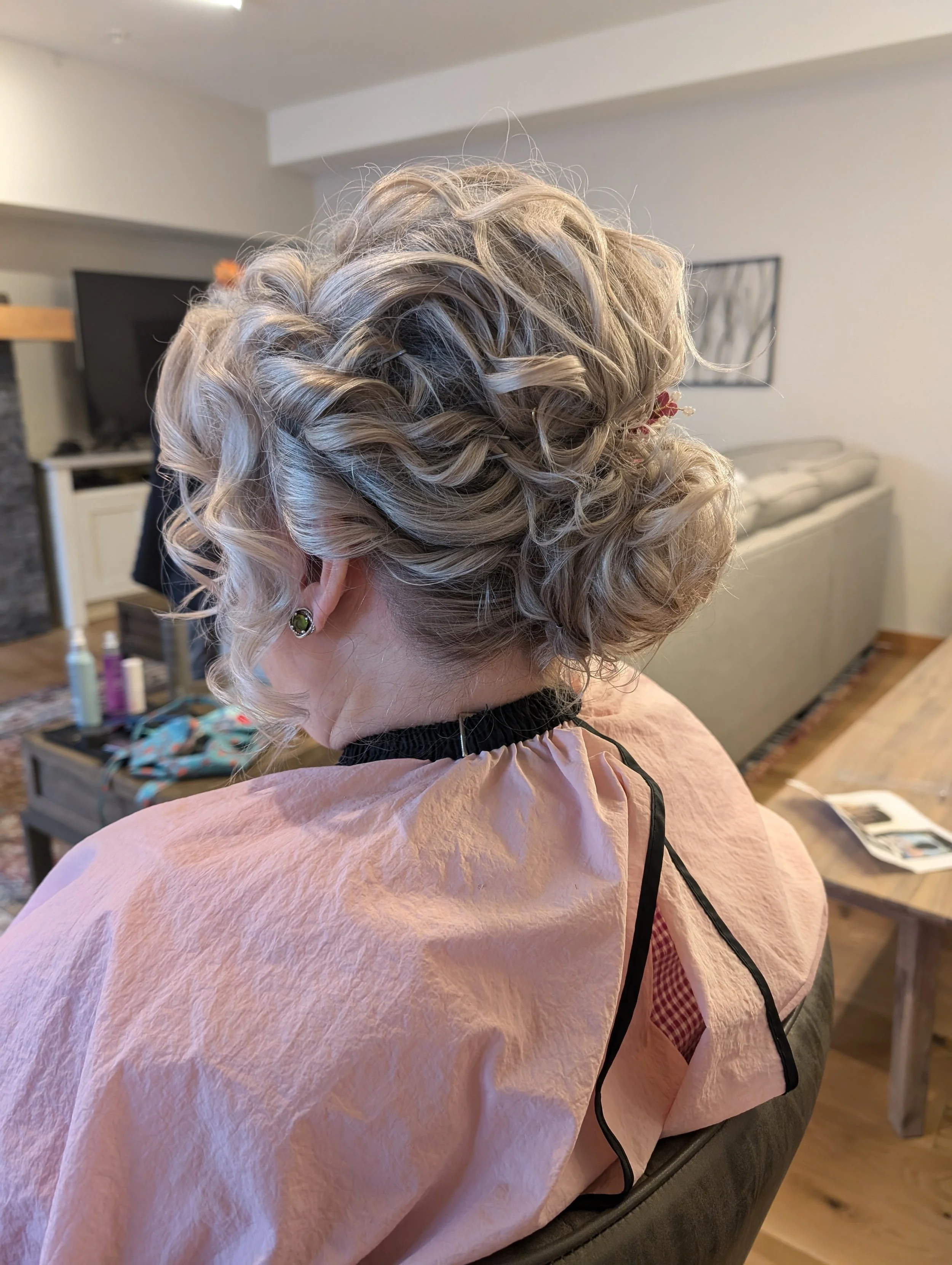 A woman with curly blonde hair styled in an elegant updo, seated indoors wearing a pink cape and a black choker.