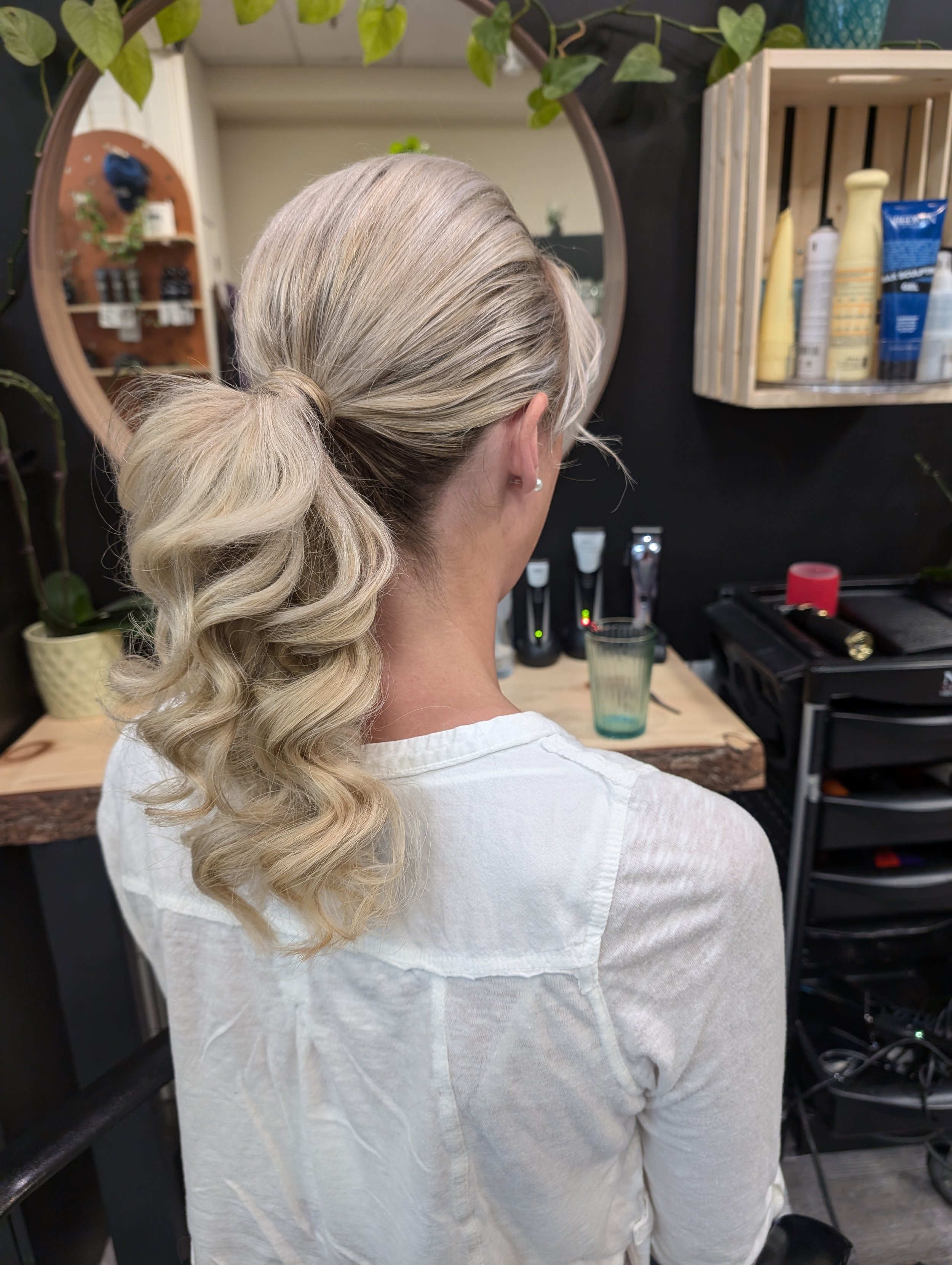 Back view of a woman with blonde, styled curly hair tied back in a ponytail, sitting in a hair salon, with a mirror, plants, and hair products visible in the background.
