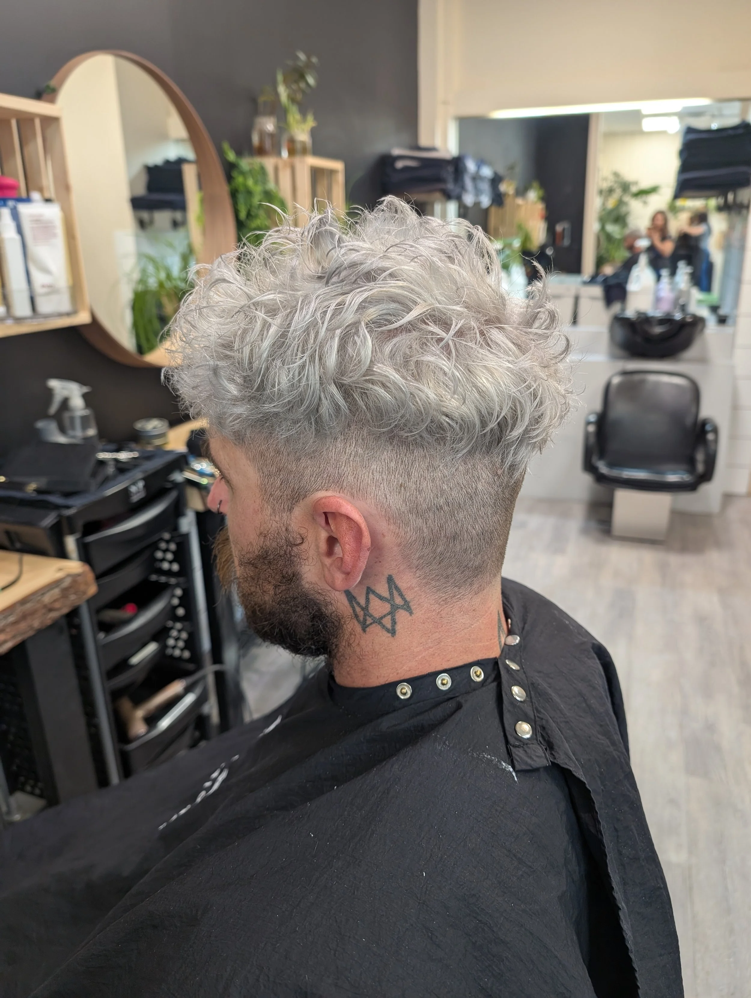 Man with curly, dyed silver hair in a salon chair, with a fade haircut and tattoo on his neck, seen from the side.