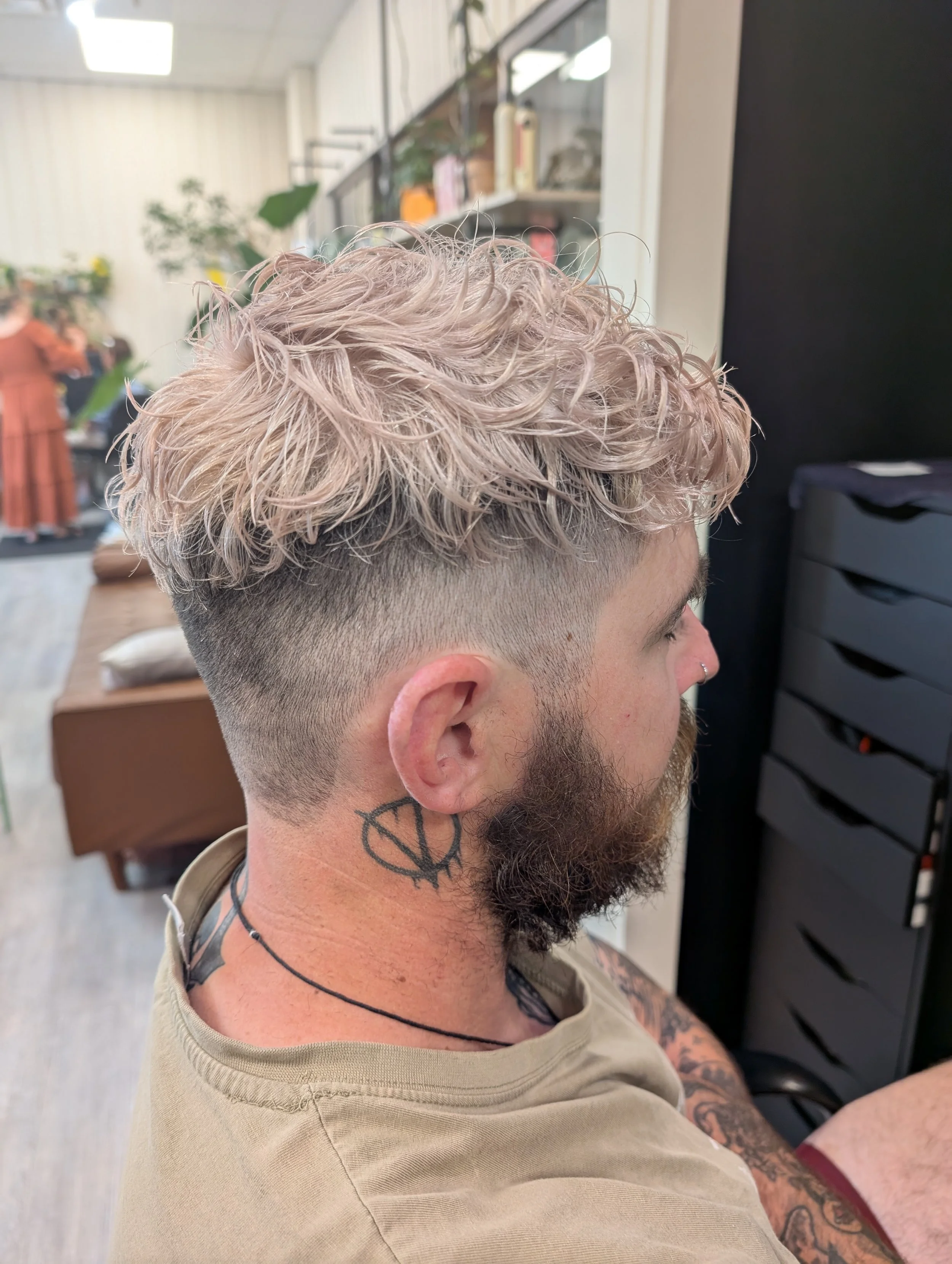 Side view of a man with a fresh haircut featuring a fade and wavy platinum blonde hair on top, tattoos on his neck and arm, and a beard, in a salon.