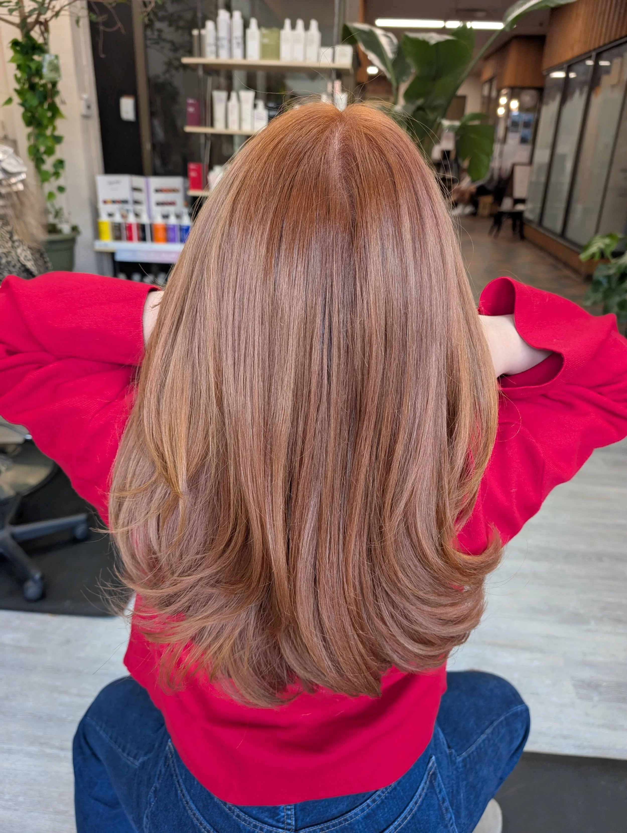 Back view of a woman with long, straight, reddish-brown hair wearing a red shirt, seated in a salon with hair products in the background.