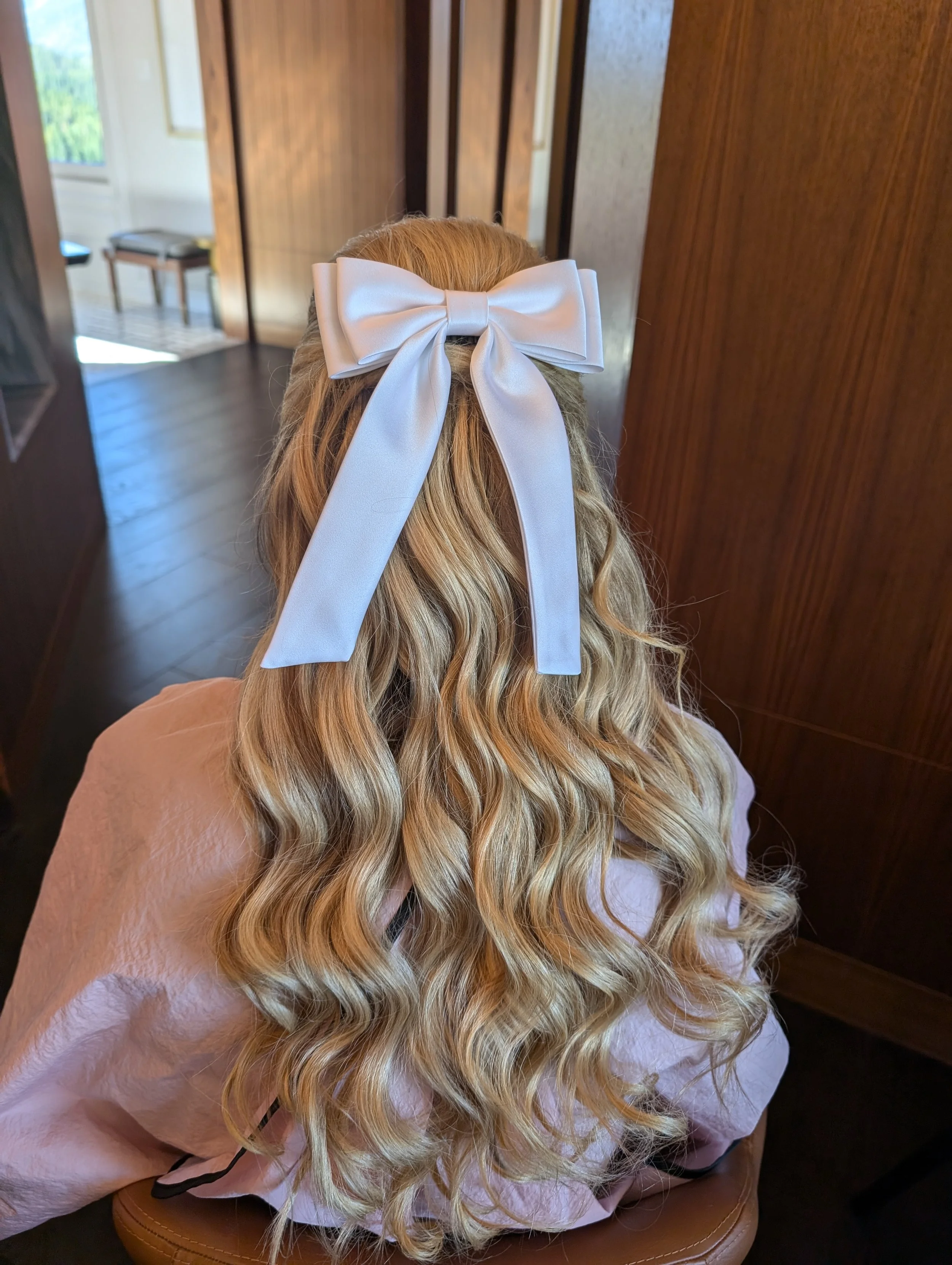 Back of a woman with long, wavy blonde hair wearing a large white satin bow in her hair, sitting indoors.