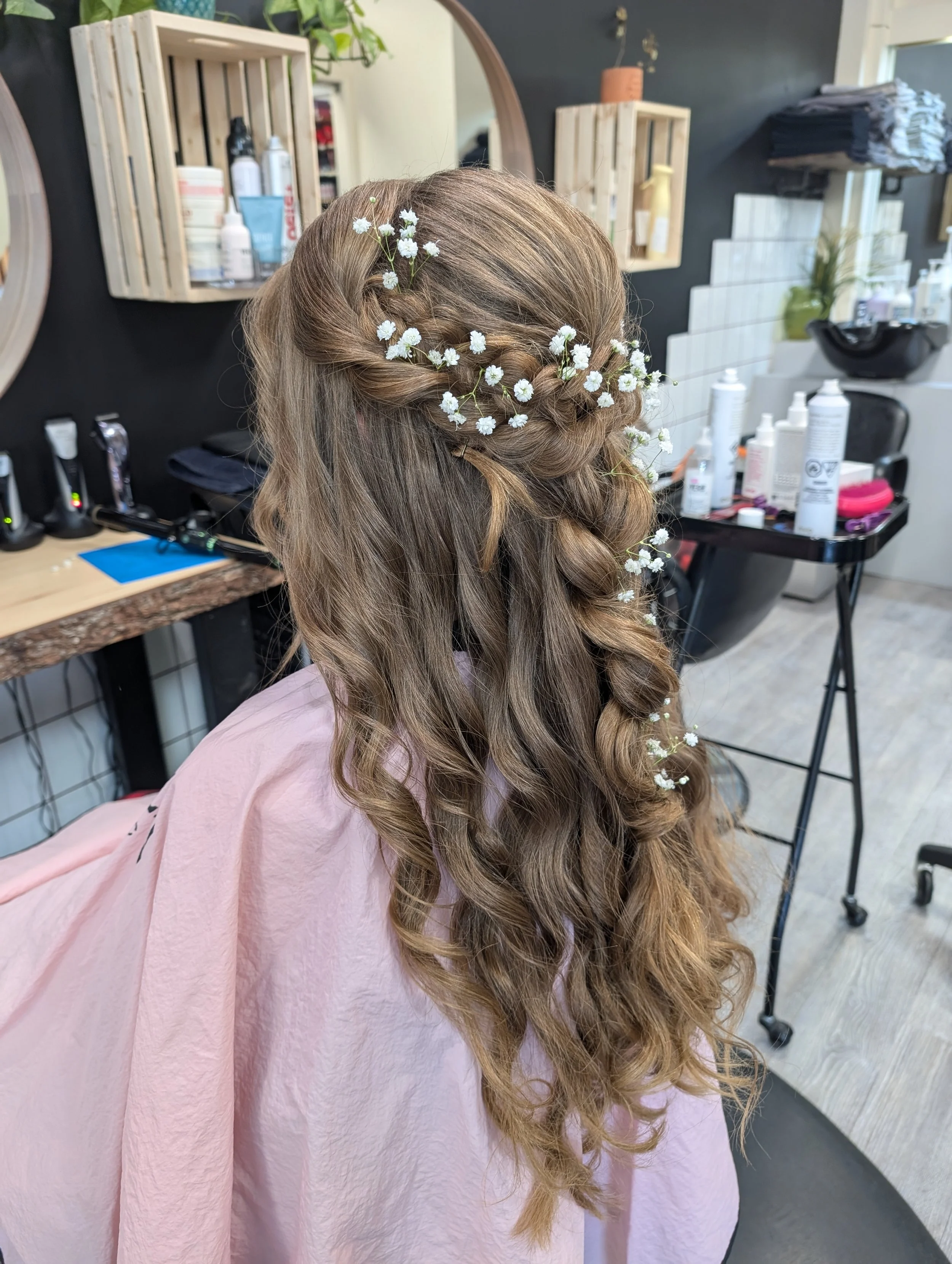 A woman with curly, styled hair decorated with small white flowers, sitting in a salon chair with a pink cape. The salon has mirrors, shelves with hair products, and styling tools visible in the background.