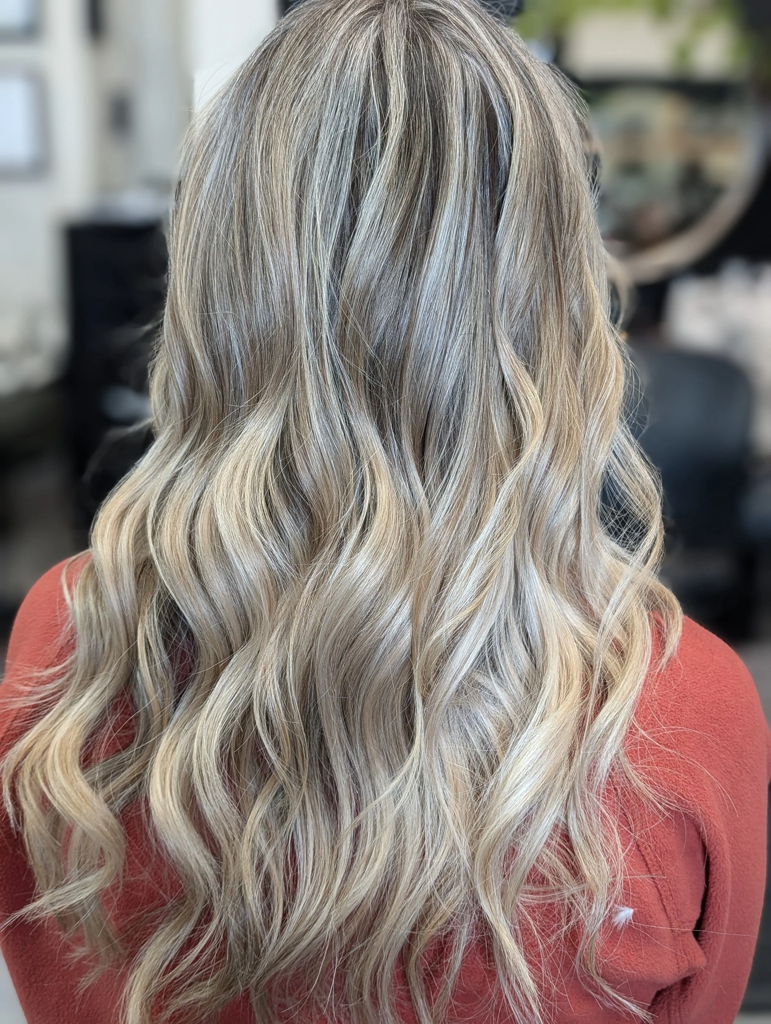 Back of a woman with long, wavy blonde hair with darker roots, wearing a rust-colored top, in a salon setting.