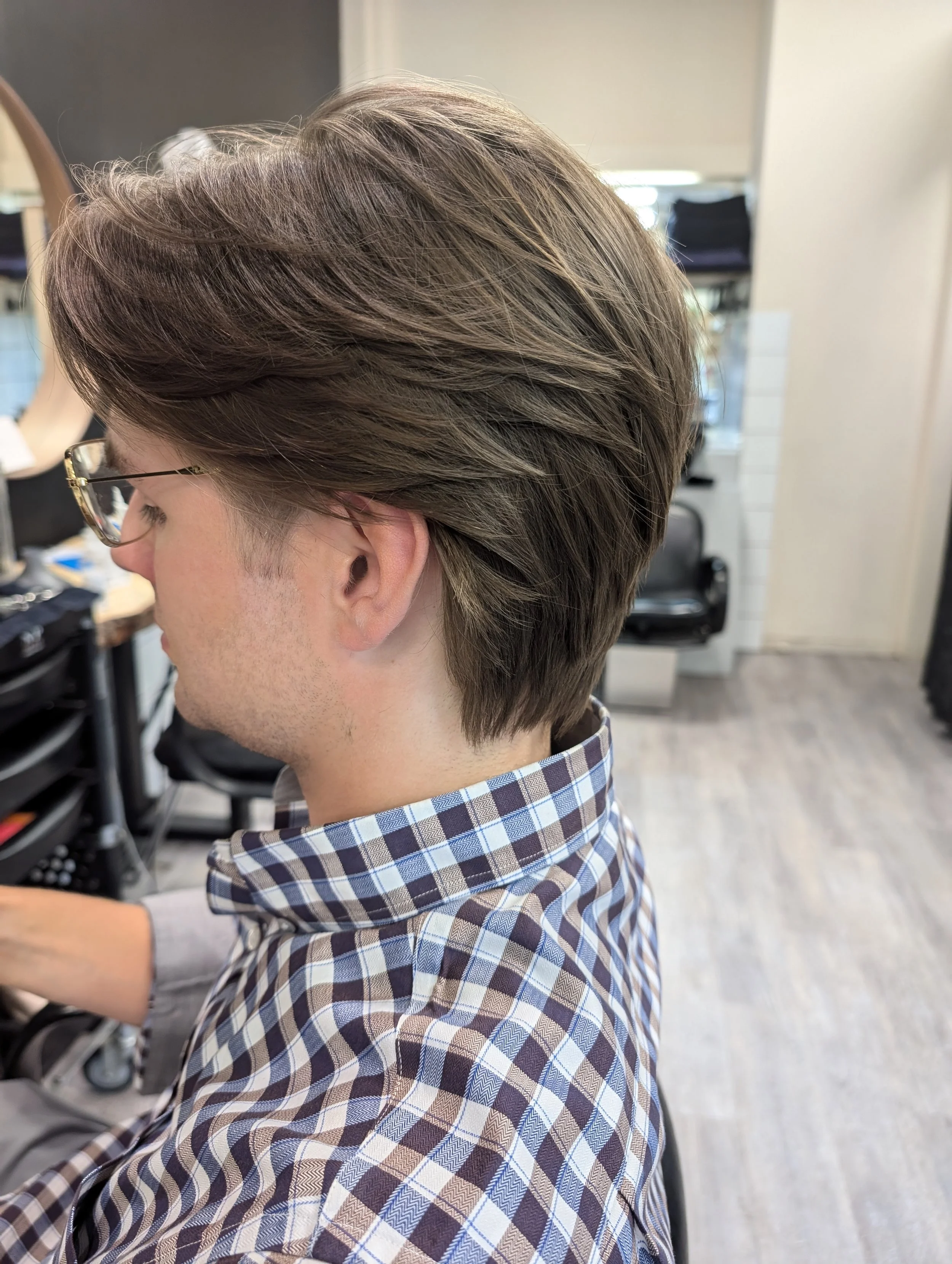 Side view of a person with layered, styled brown hair and glasses, wearing a checkered shirt, sitting in a salon or barber shop.