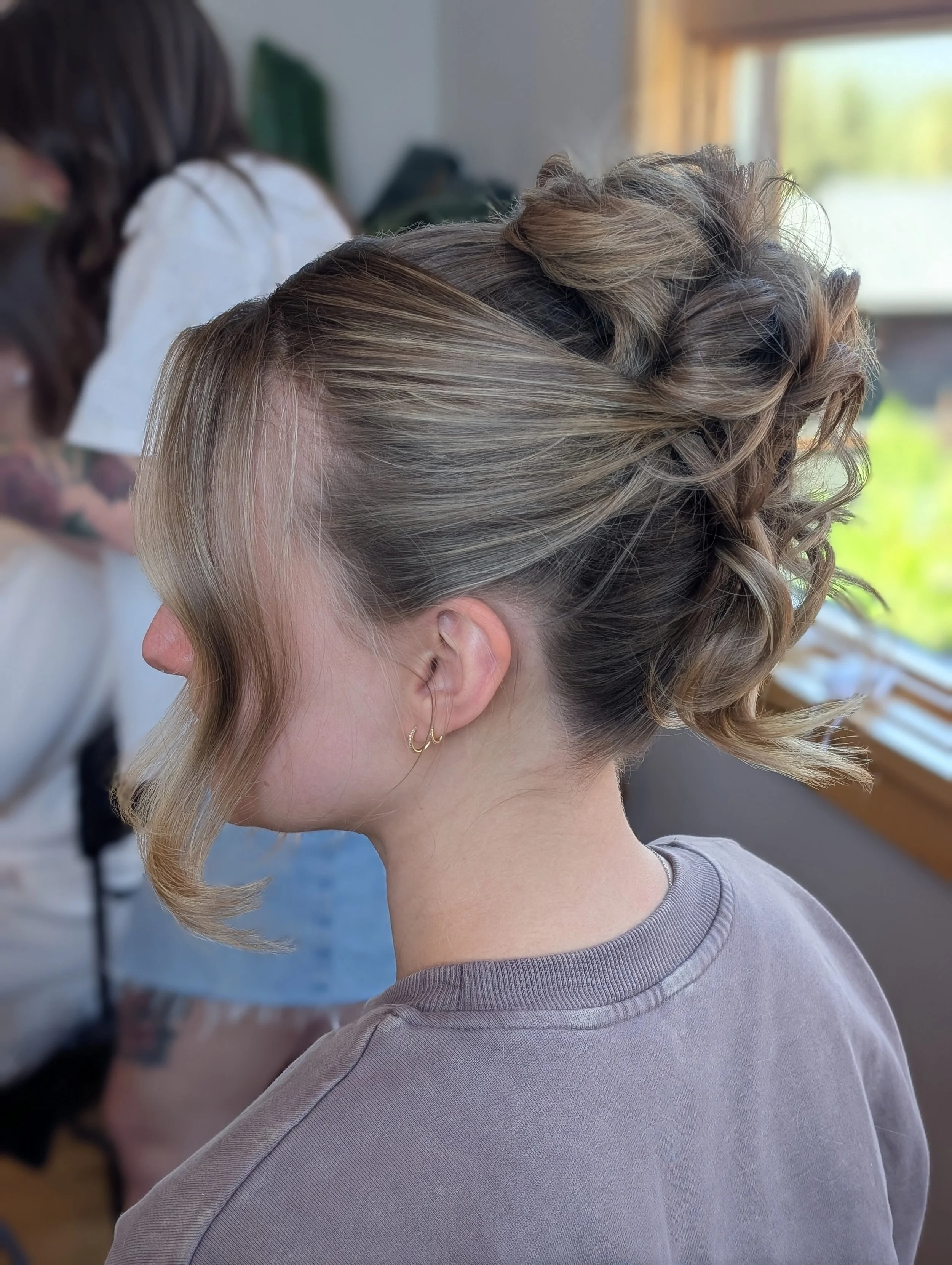 A woman with blonde hair styled in an elegant updo with curls, wearing small gold hoop earrings and a gray shirt, in a bright room with a window.