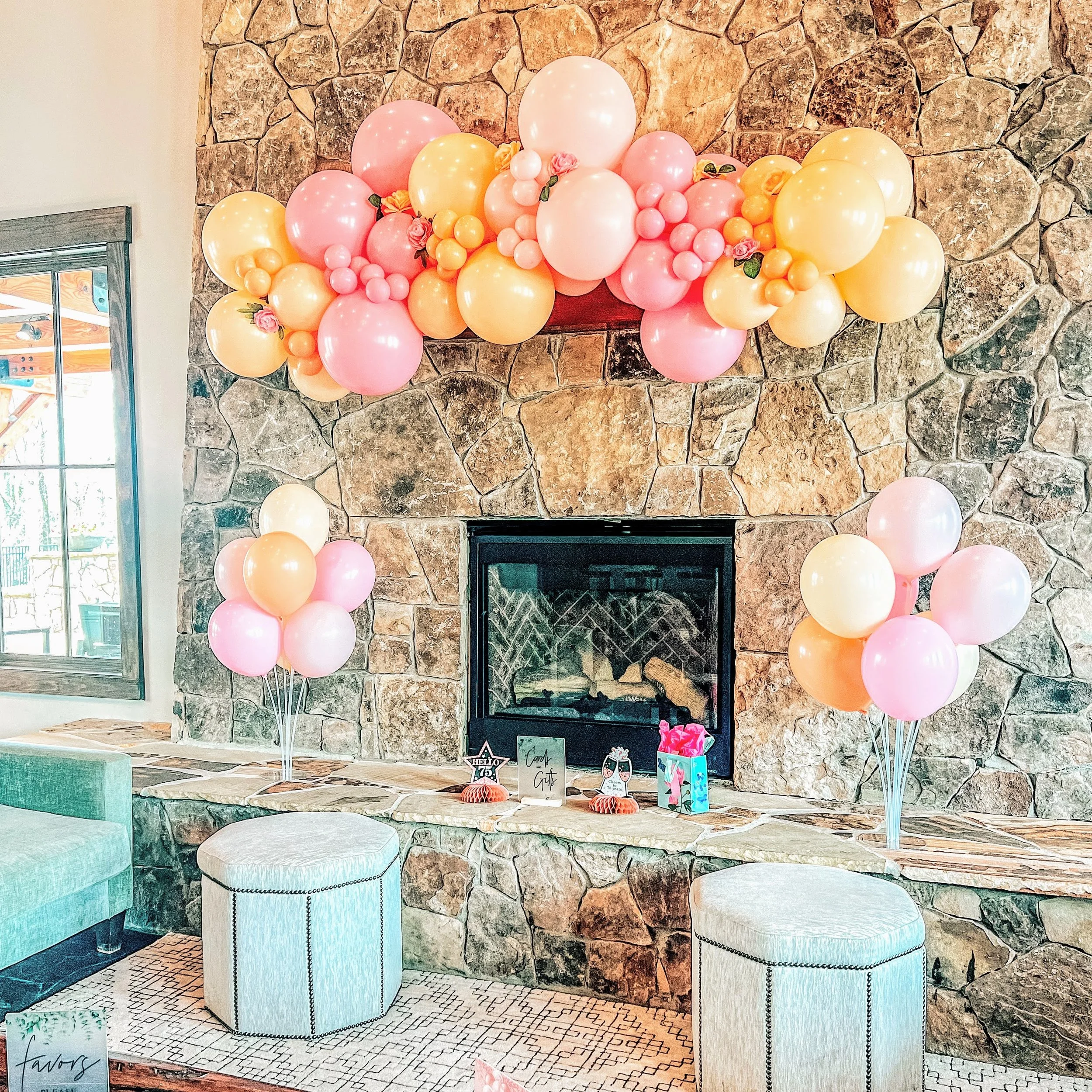 Decorative pink, yellow, and peach balloons arranged around a stone fireplace with small party decorations on the hearth, including gift boxes, framed photos, and a sign that says "Girl".