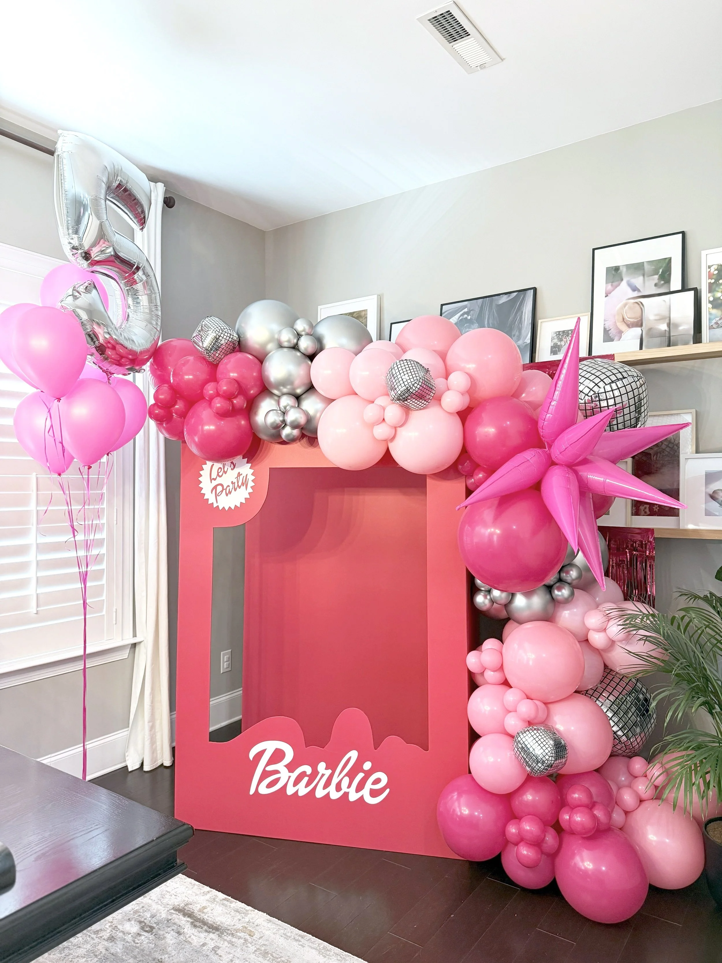 Pink Barbie-themed photo booth with pink, silver, and black balloons, a large pink frame with the Barbie logo, and pink balloons shaped like stars at a birthday party.