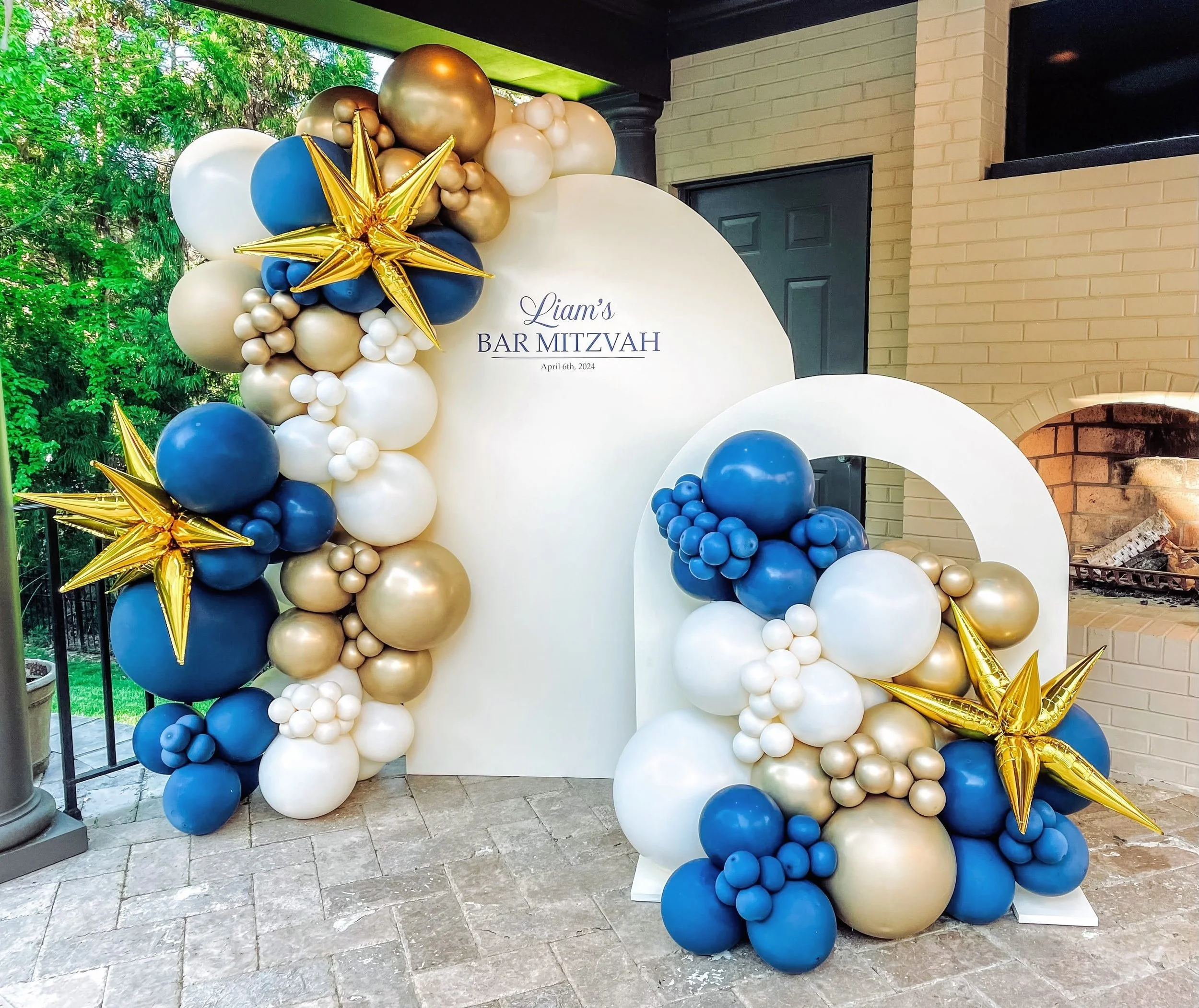 Balloon arch with gold, white, and navy blue balloons, gold star-shaped balloons, and a sign reading "Liam's Bar Mitzvah, April 6th, 2024" on a white backdrop.