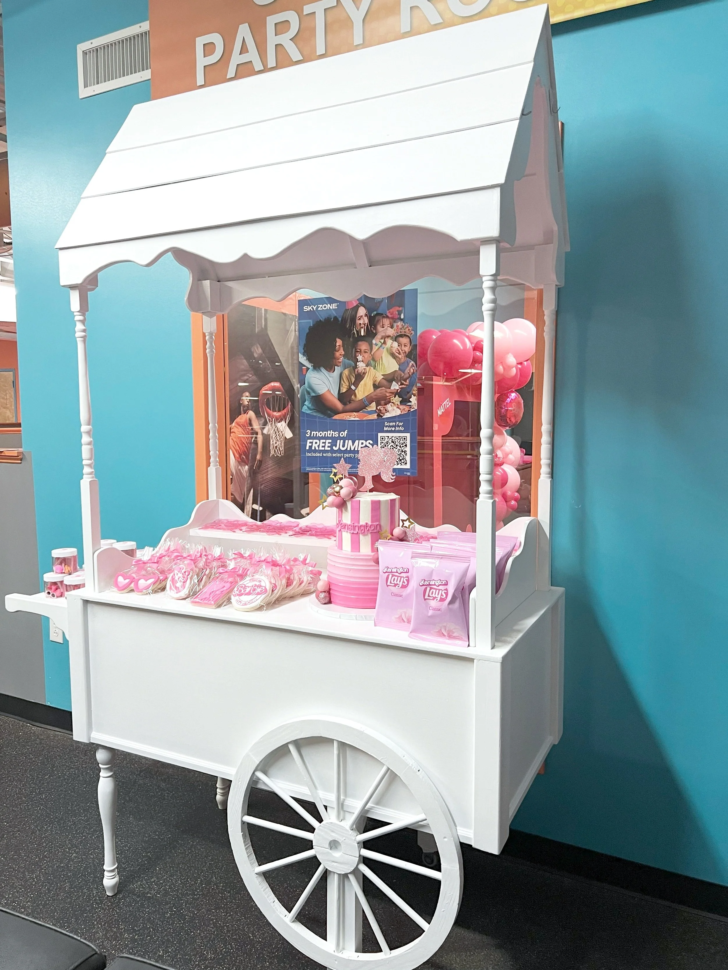 Pink and white dessert stand cart with a striped cake, pink balloons, and pink candy bags, against a blue wall.