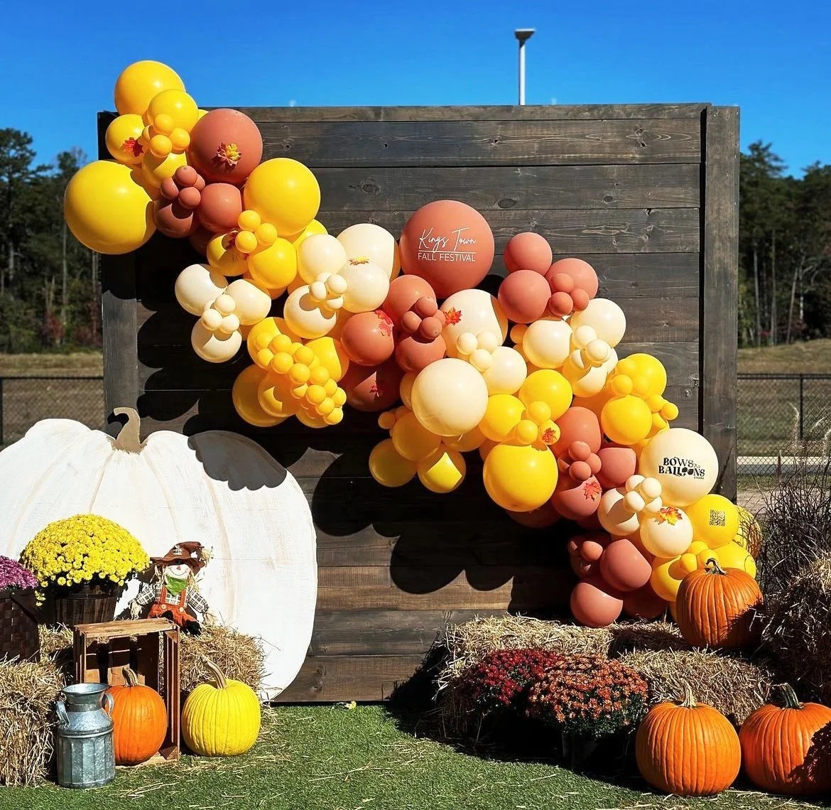 Fall festival display with a wooden backdrop decorated with a large cluster of yellow, orange, and cream balloons, pumpkins, hay bales, potted chrysanthemums, and a large white pumpkin with autumn decor.