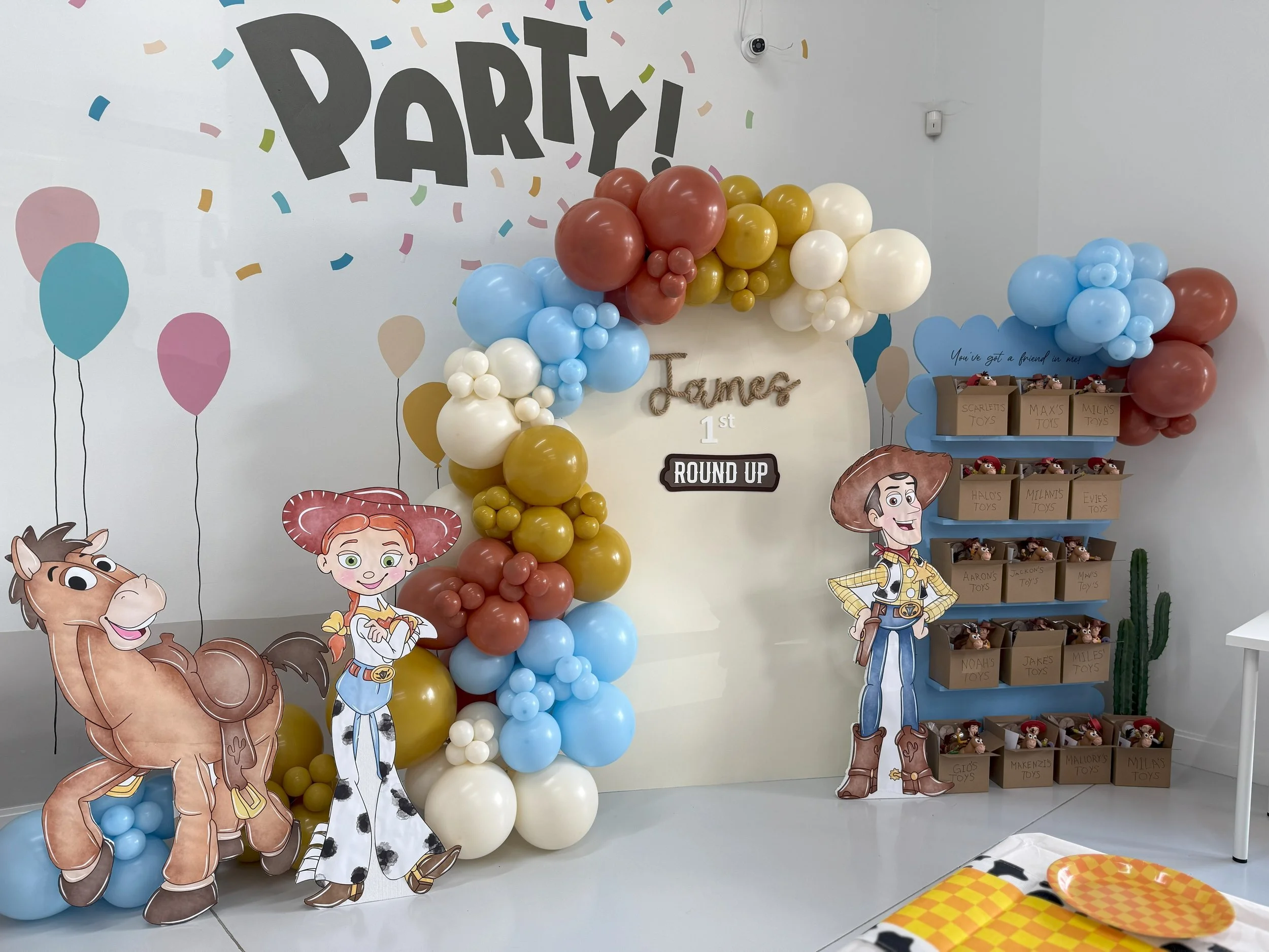 Decorated party space with cartoon Toy Story characters Woody, Jessie, and Bullseye, colorful balloon arch, and storage boxes of toys.