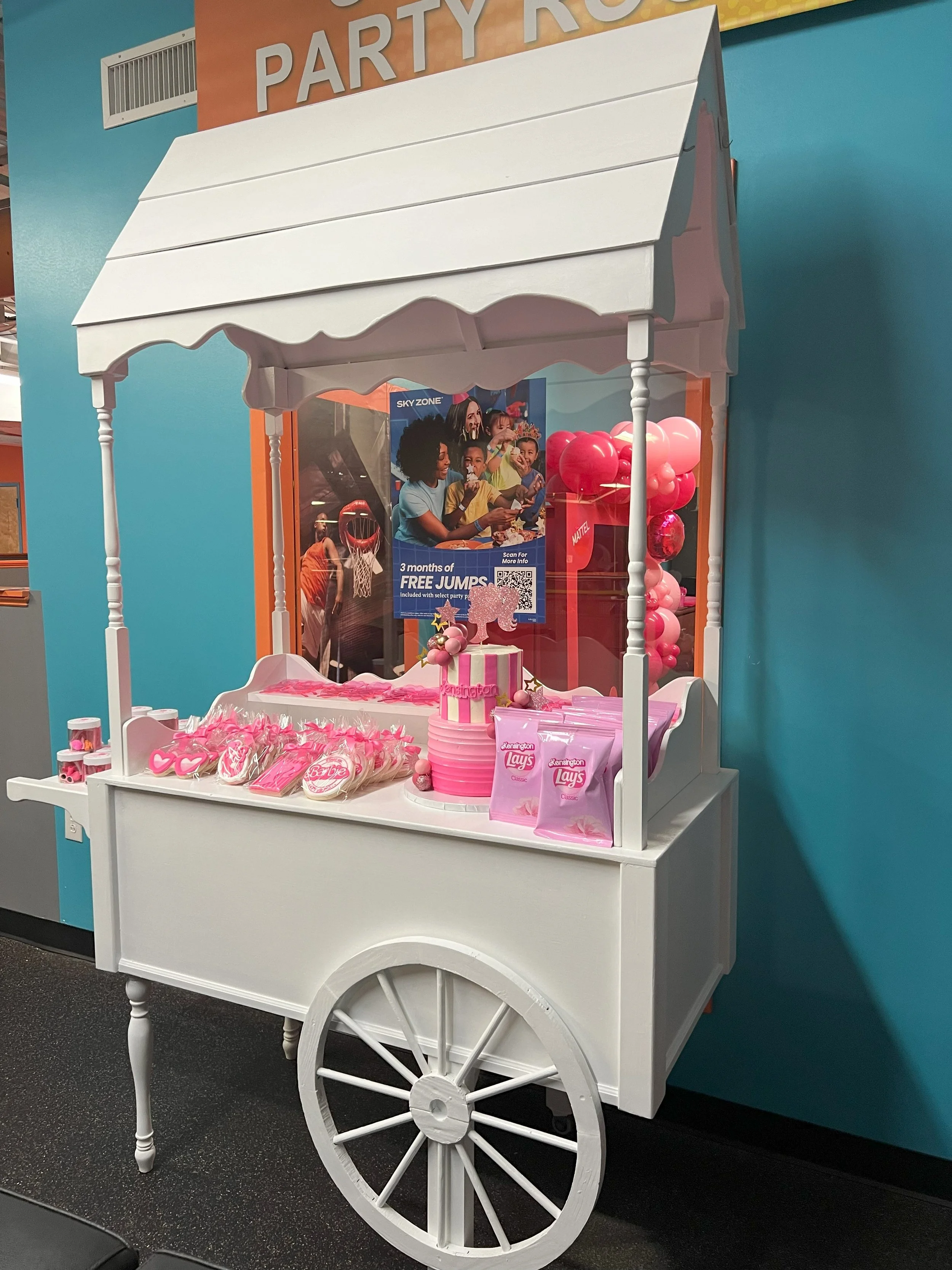 A pink and white party cart decorated with candies, cupcakes, and balloons, with a pink striped cake topped with a pink glittery unicorn, set against a teal wall with an orange banner overhead.