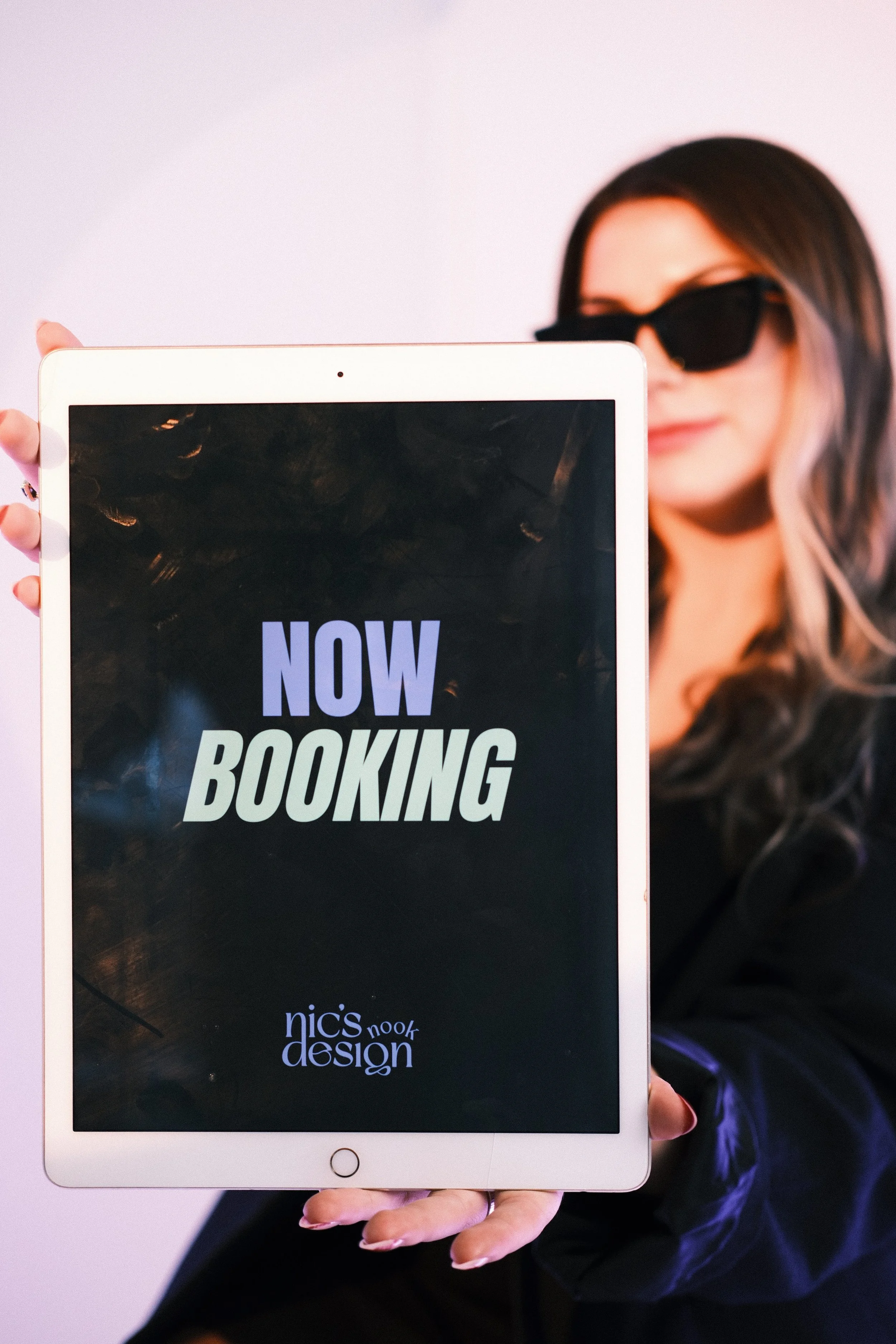A woman with dark hair wearing black sunglasses holds a tablet screen displaying 'NOW BOOKING' in colorful letters, with a white background.