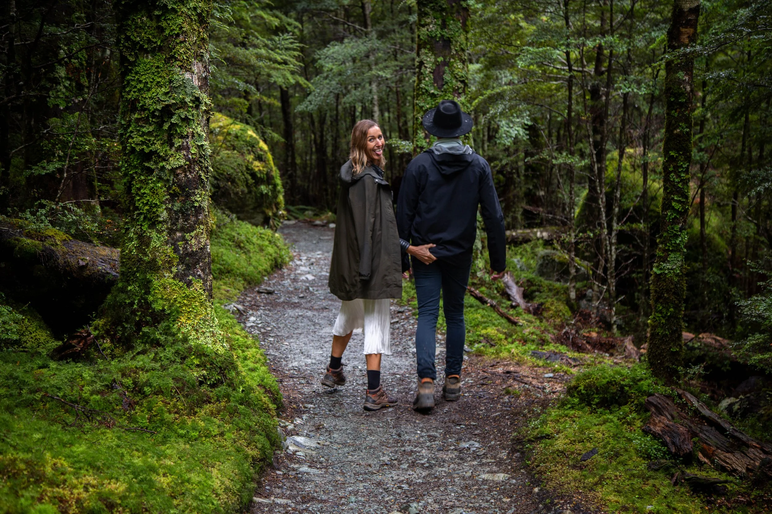 Two women holding hands while walking on a trail through a dense, green forest.