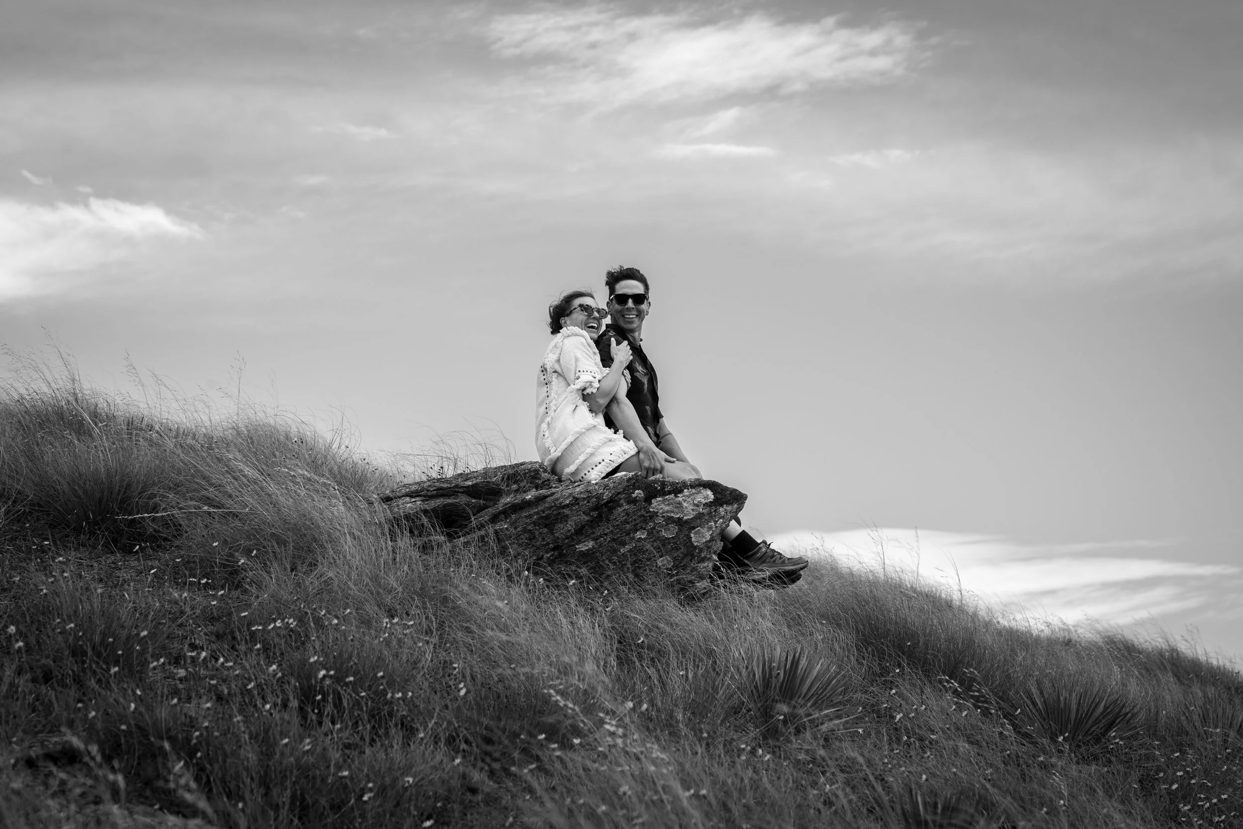 A black and white photo of two people sitting on a large rock on a grassy hill, smiling and laughing, with a woman wearing glasses and a light-colored sweater and a man wearing sunglasses and a dark shirt.