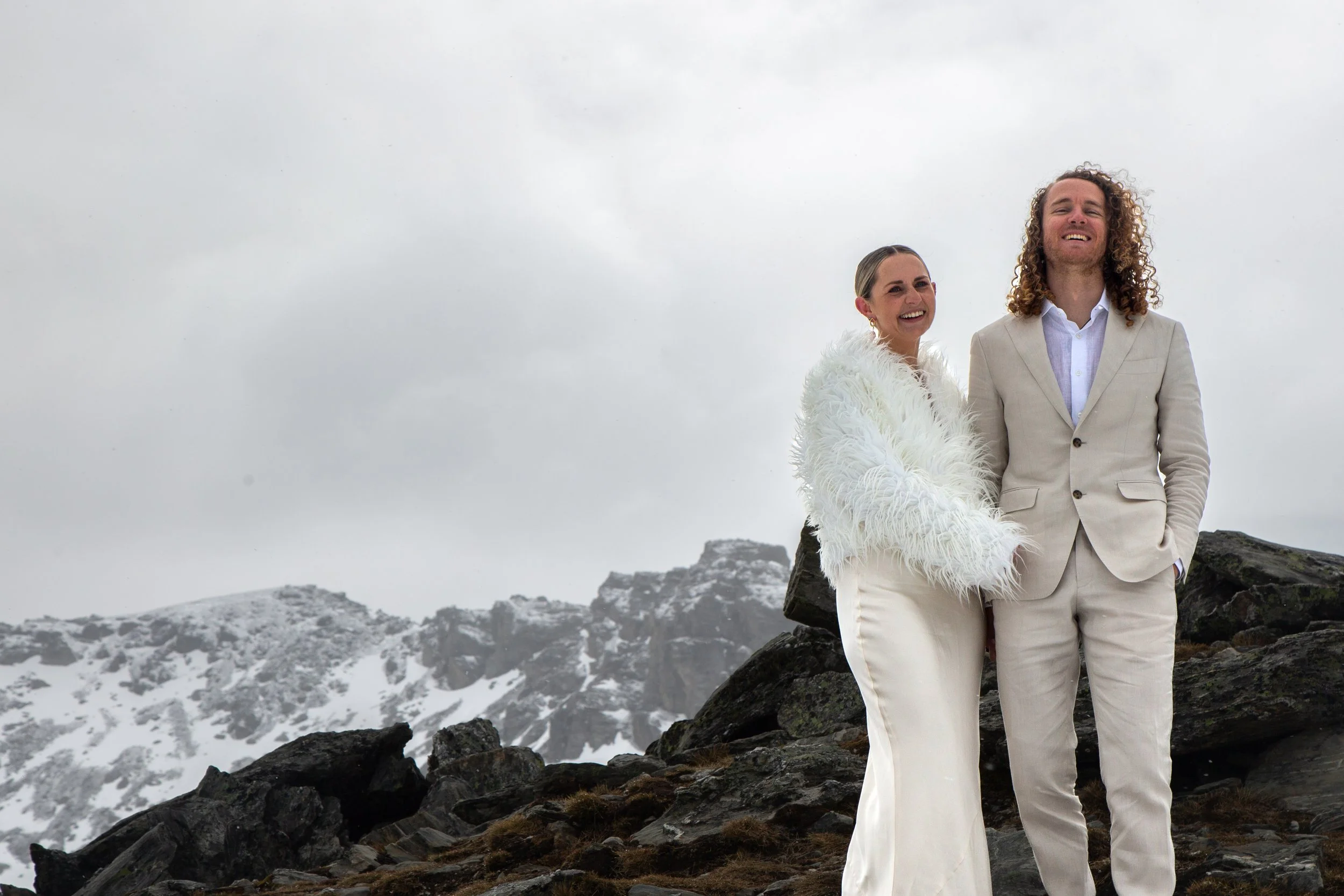 A happy woman and man standing outdoors on rocky terrain with snow-covered mountains in the background. The woman is wearing a white furry jacket and white pants, and the man is wearing a light beige suit with a white shirt. They are smiling and enjo