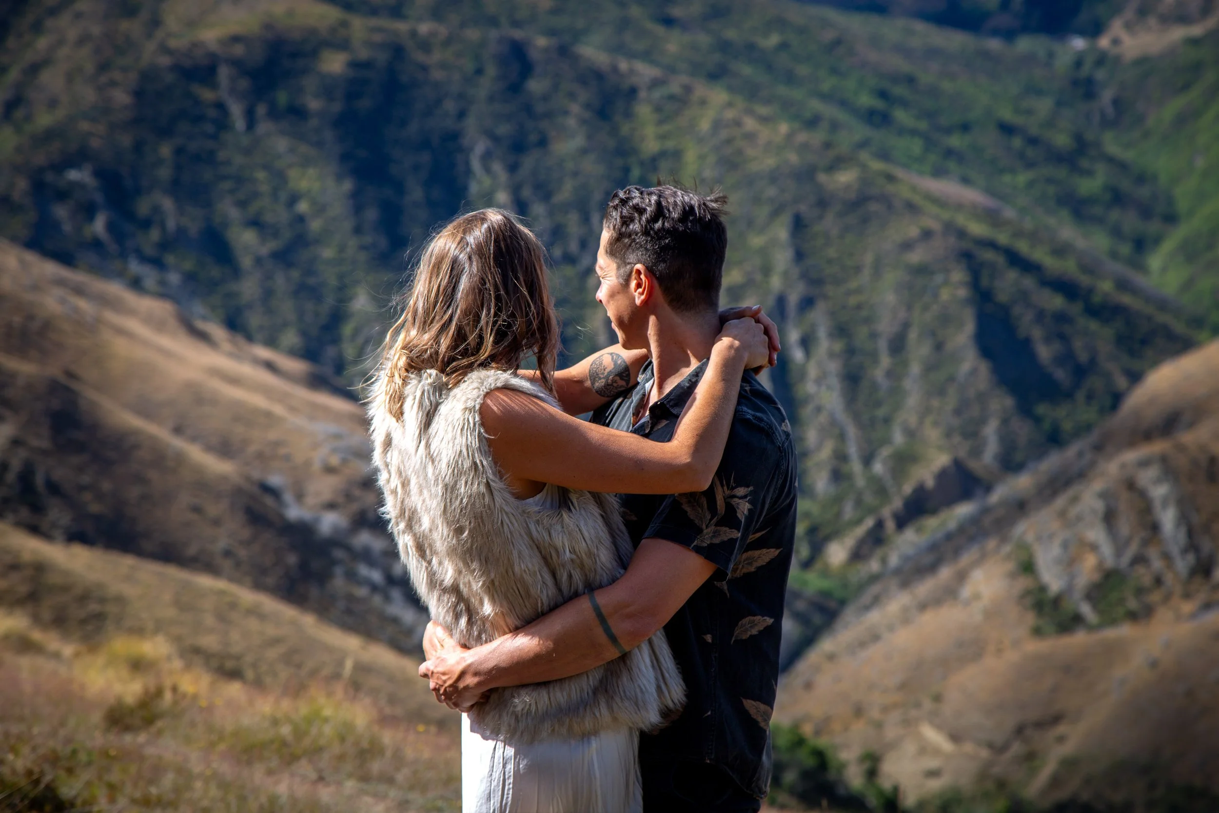 A couple holding each other and smiling in a mountainous outdoor setting.