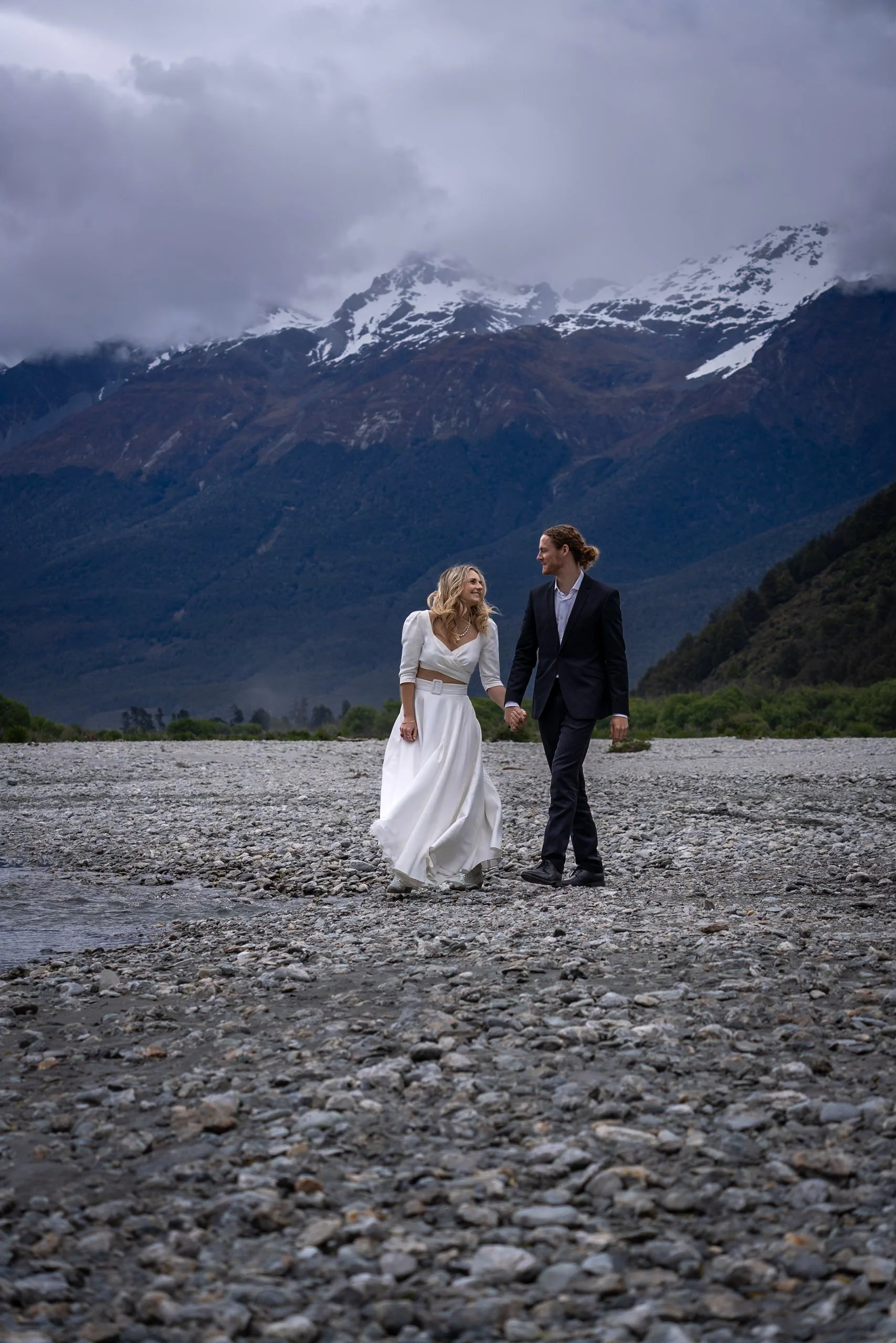 A couple in wedding attire holding hands and walking on a rocky riverbank with mountains in the background.