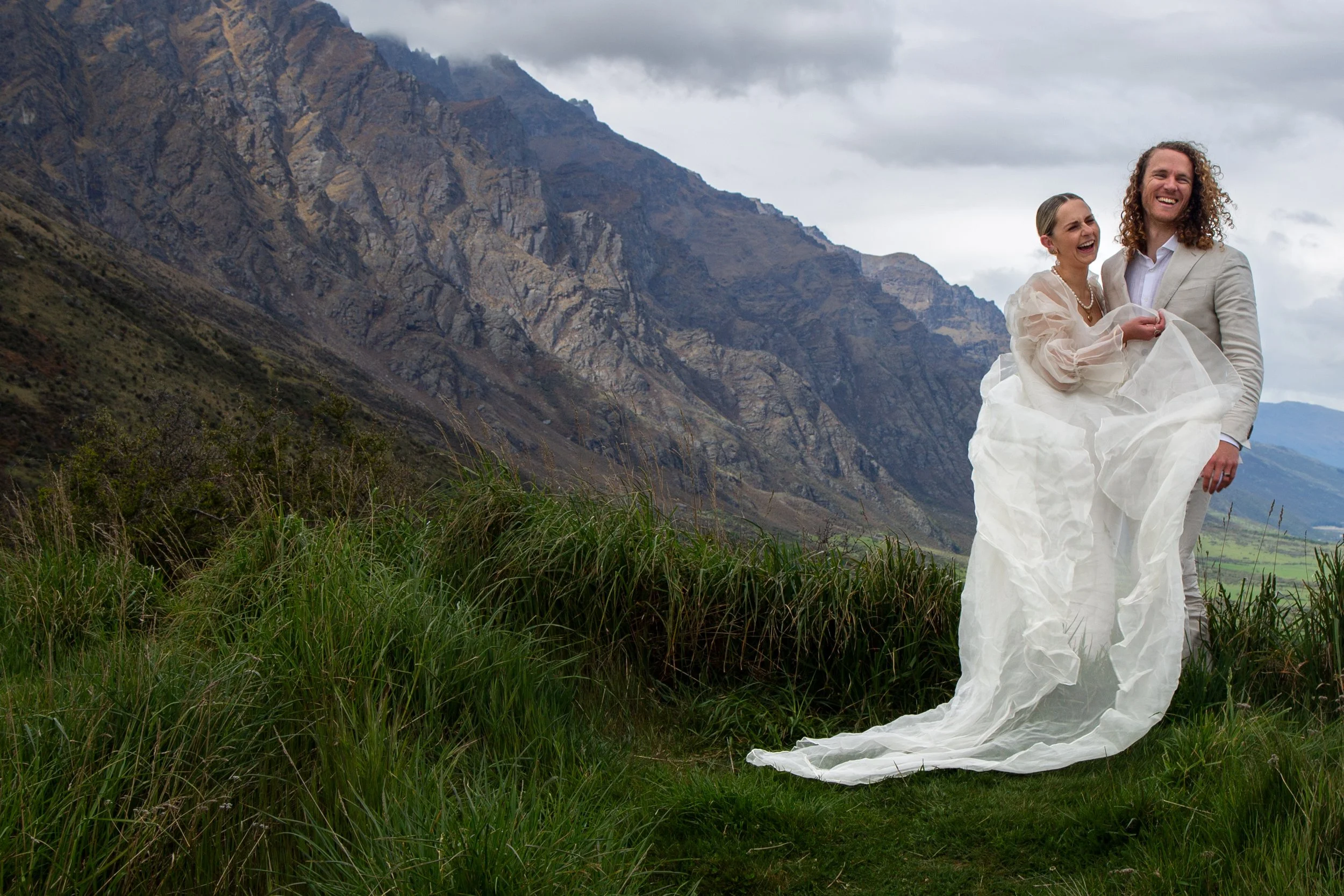 A couple in wedding attire smiling and laughing outdoors with a mountainous landscape in the background, the bride holding her wedding dress.