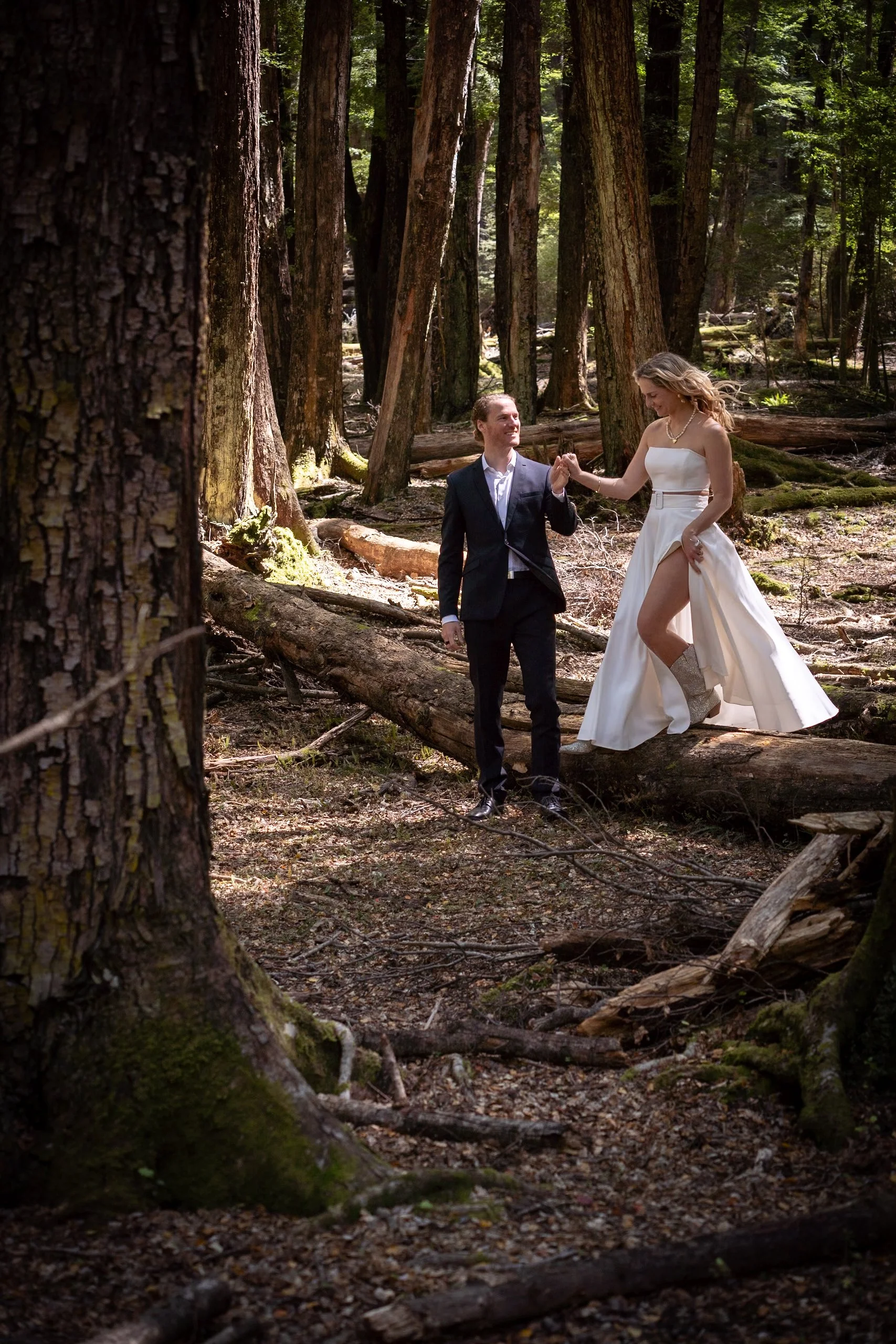 A bride and groom in wedding attire are dancing in a forest among tall trees, with the bride lifting her dress to reveal her boots.