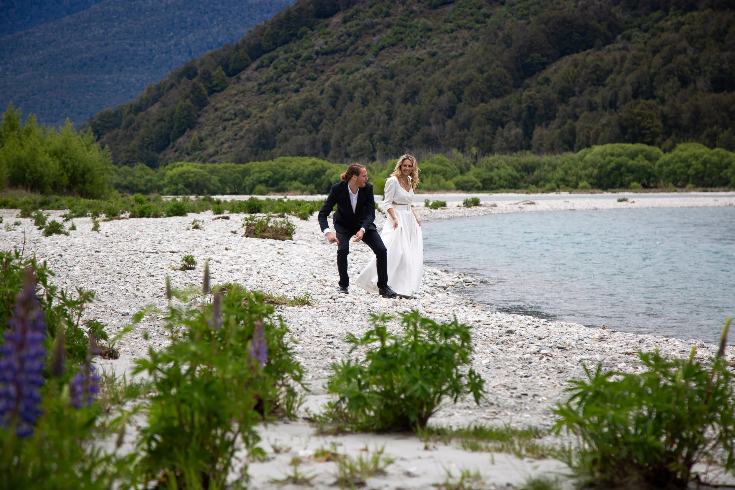A bride and groom walk along a rocky lakeshore with mountains and lush green trees in the background.