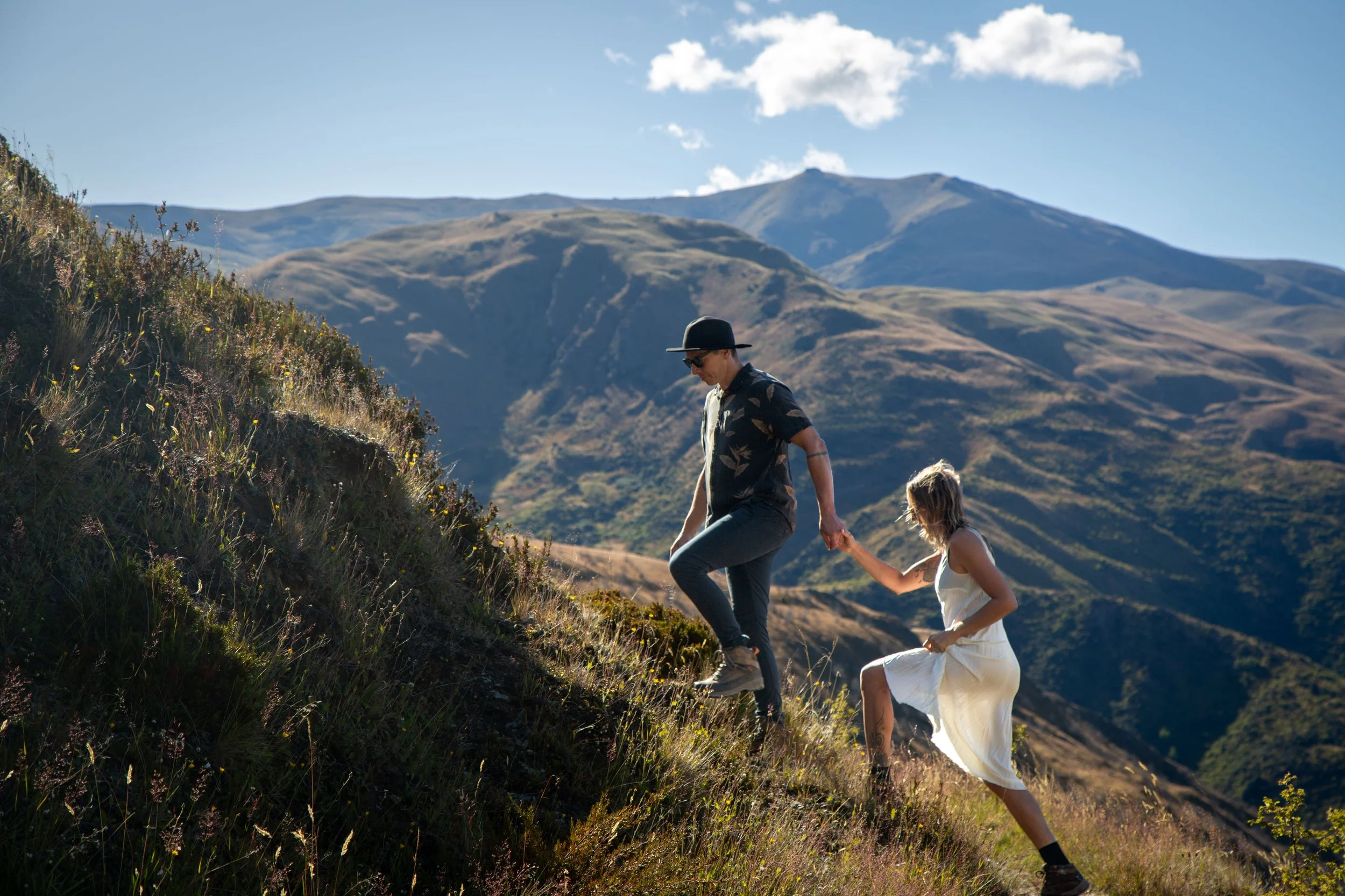 A man and woman hiking up a mountain trail in a scenic outdoor landscape with hills and mountains in the background during daytime.