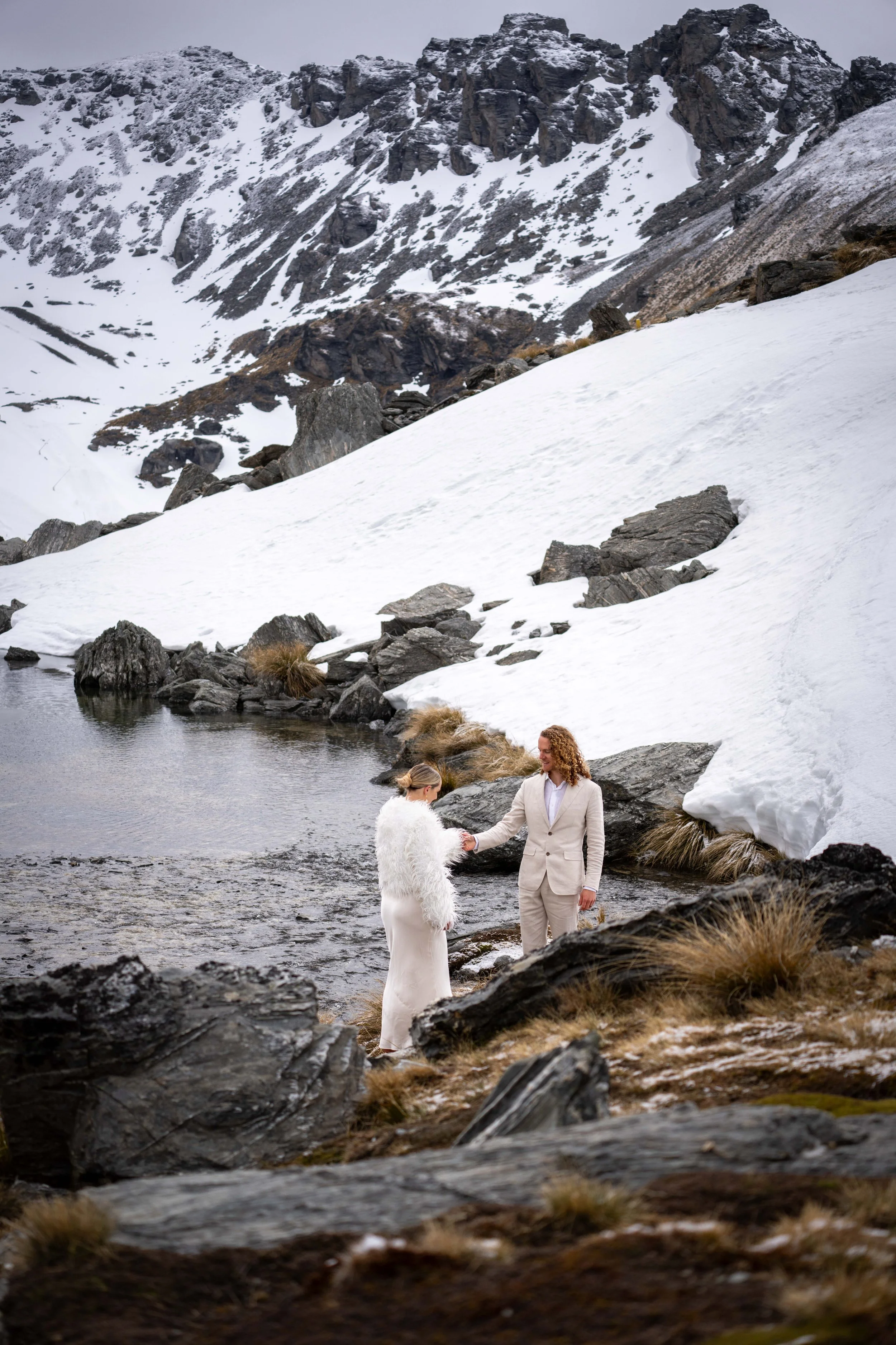 A couple dressed in light-colored clothing, standing by a mountain lake with snow-covered peaks and rocks in the background, with the man taking the woman's hand.