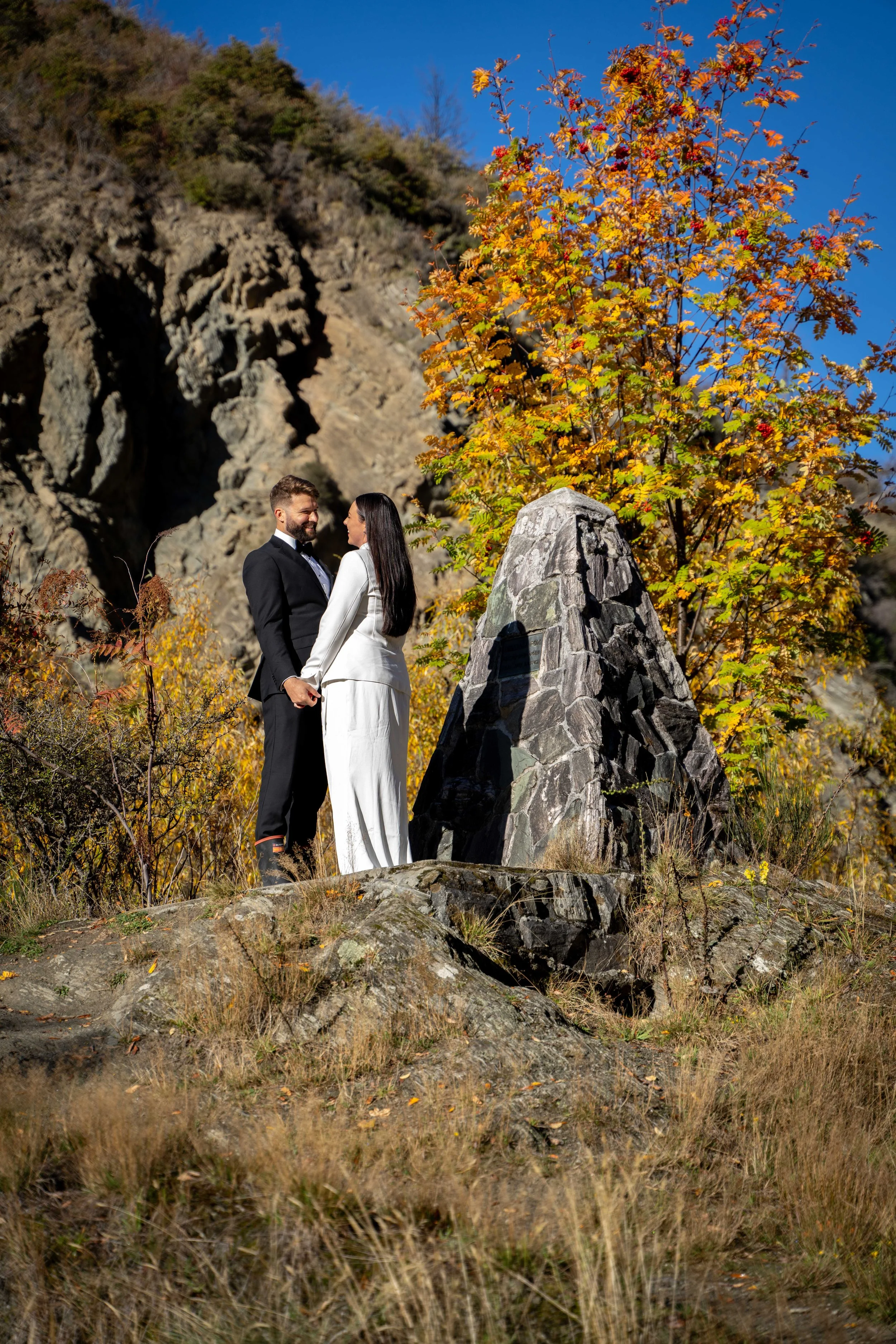A couple in wedding attire holding hands and smiling at each other outdoors in autumn, standing on a rocky area near a large rock formation and colorful orange and yellow trees.