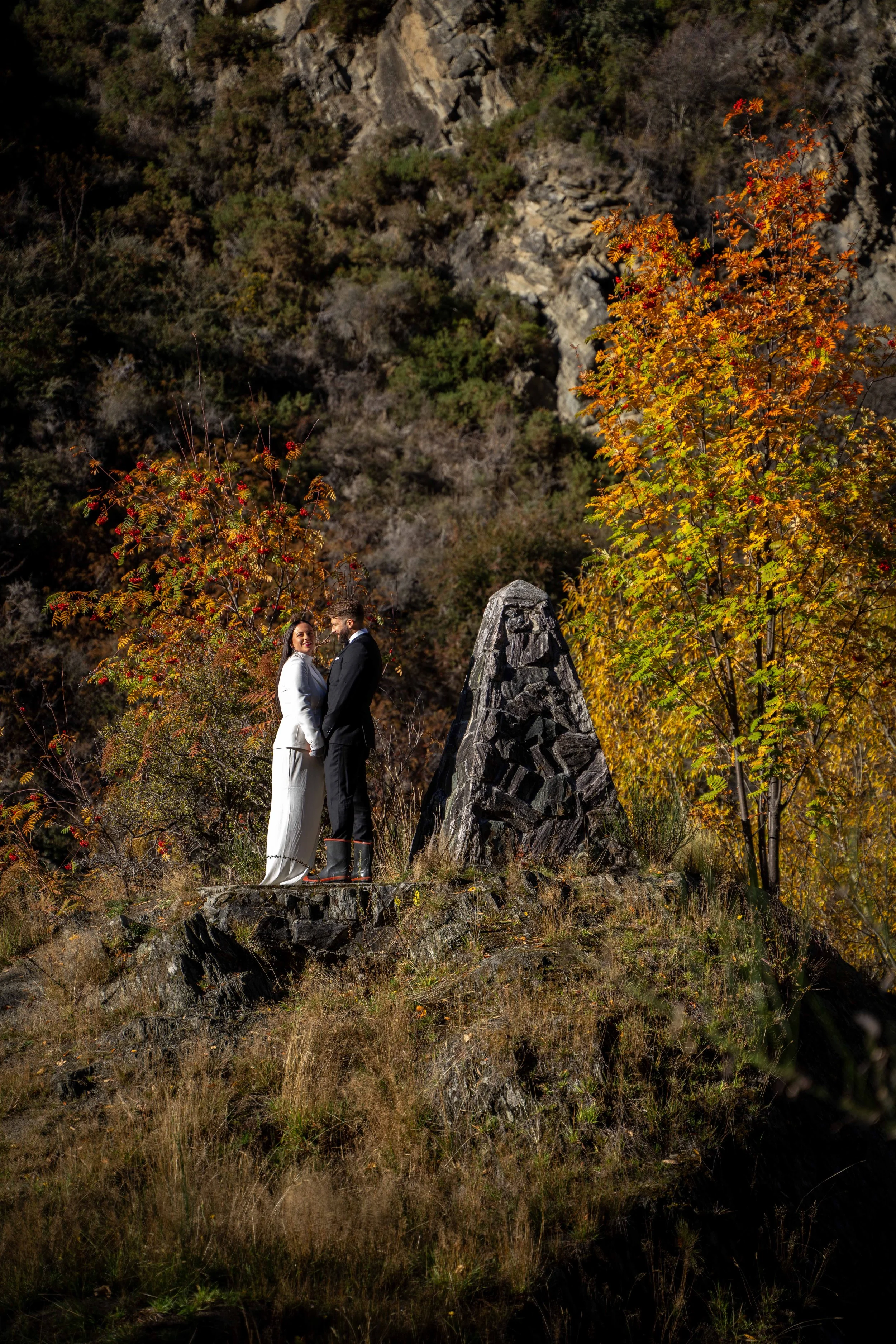 A bride and groom dressed in wedding attire stand on a rocky hill with autumn foliage and trees around them, holding hands and smiling during daylight.