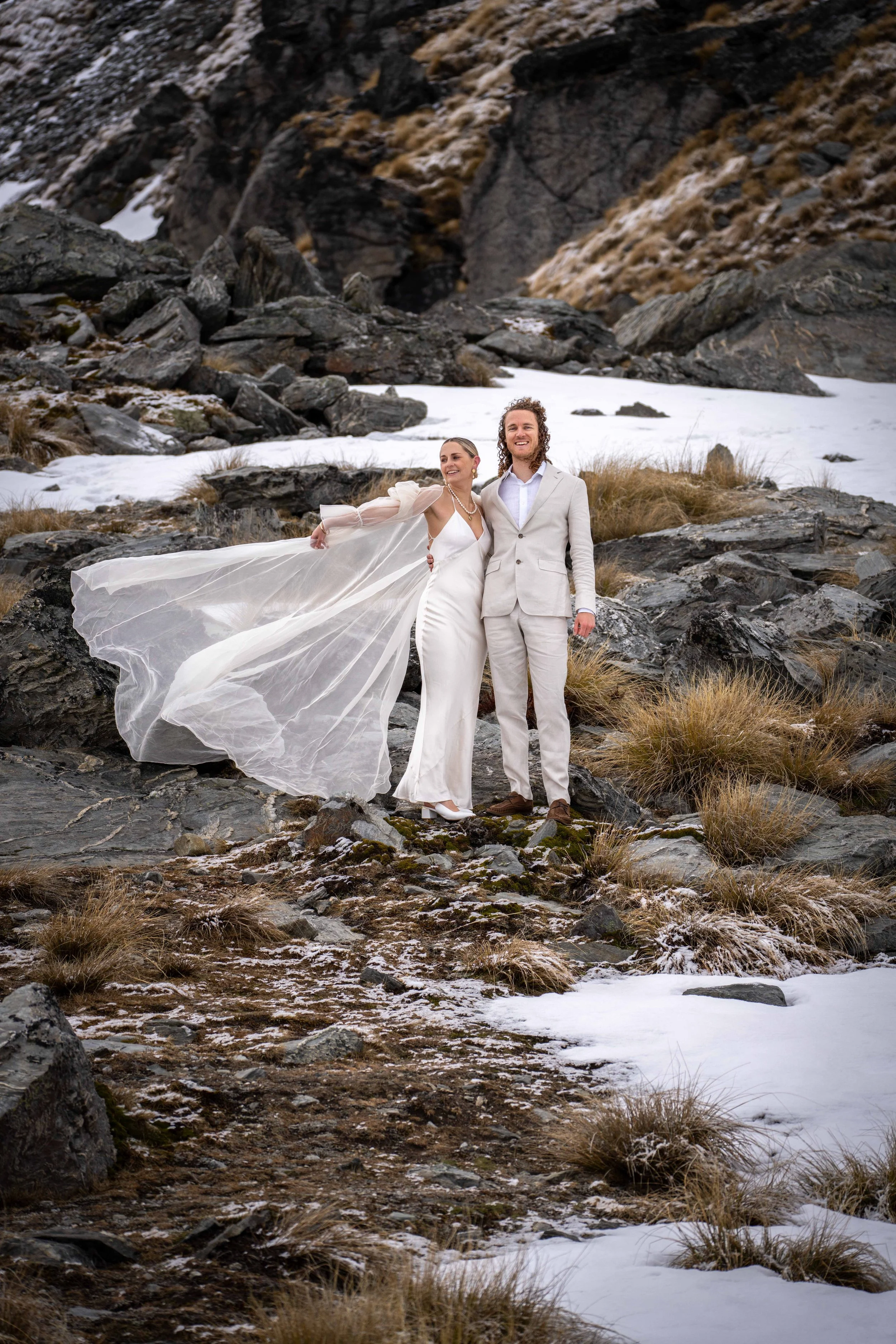 A bride and groom standing on rocky terrain with patches of snow, surrounded by mountains and dry grass, smiling after a wedding.