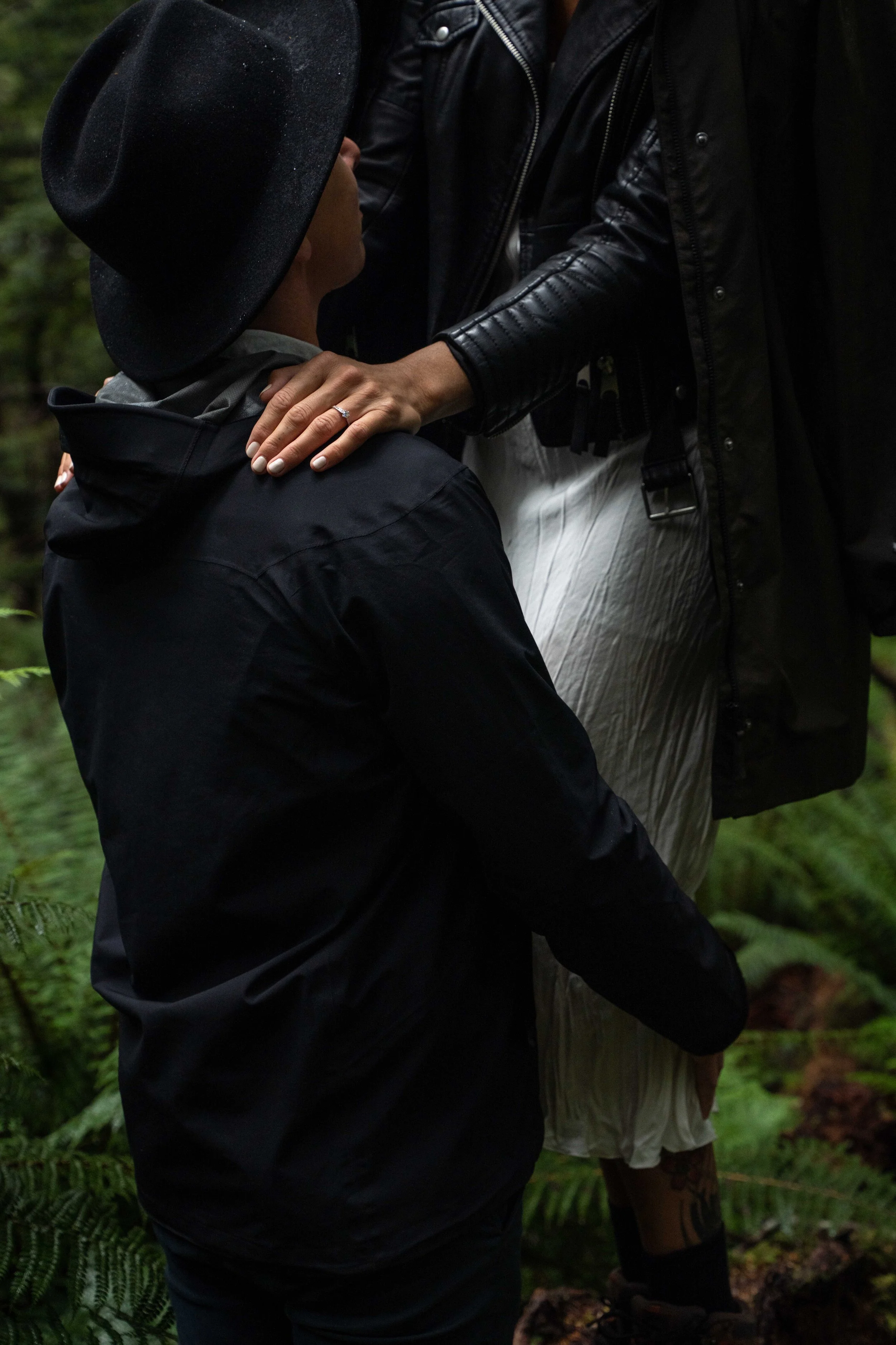 A couple in a forest, with the woman touching the man's shoulder, the woman wearing a white dress and the man in a black jacket and hat.