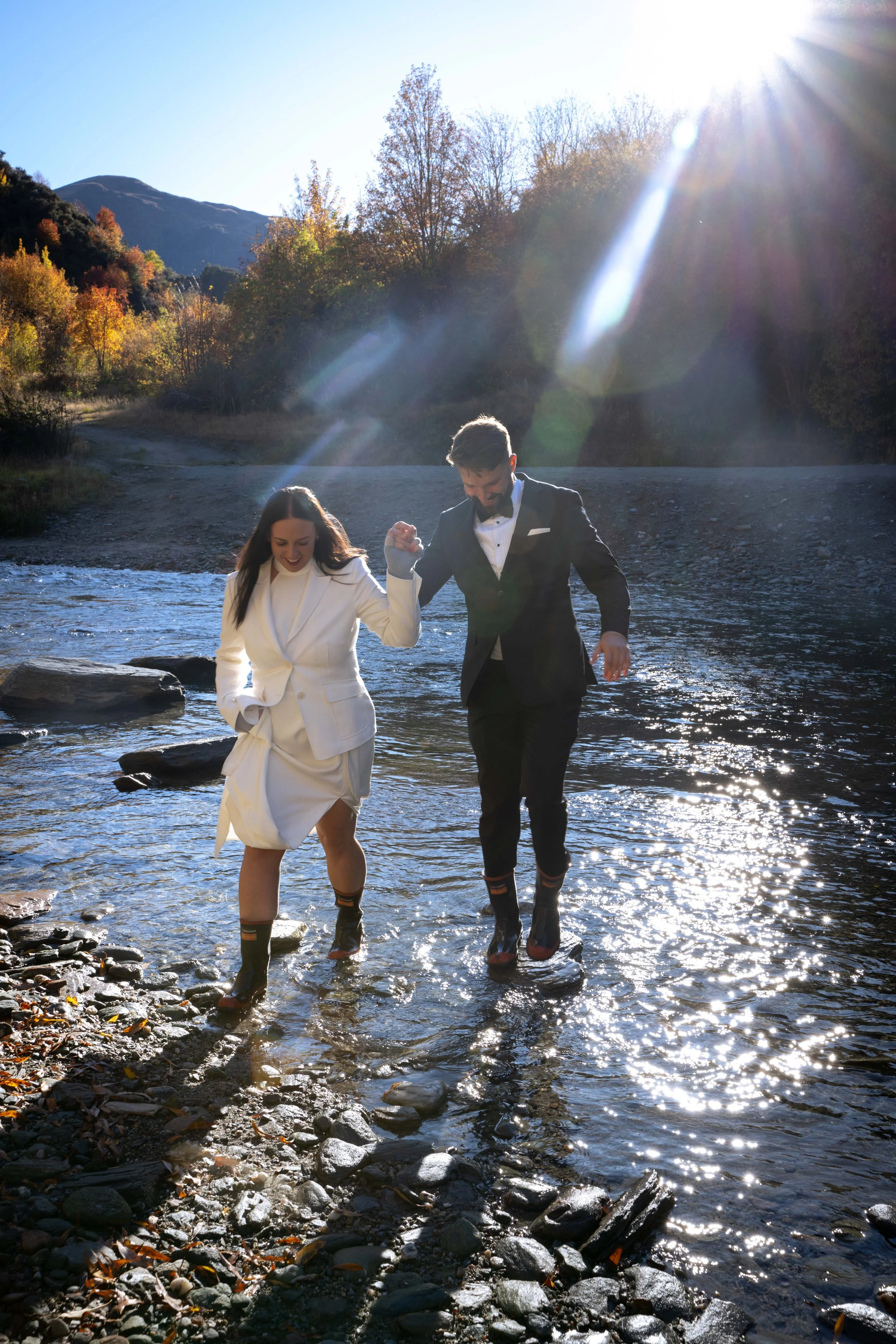A man and woman dressed in formal attire crossing a rocky stream with the sun shining behind them and trees in the background.