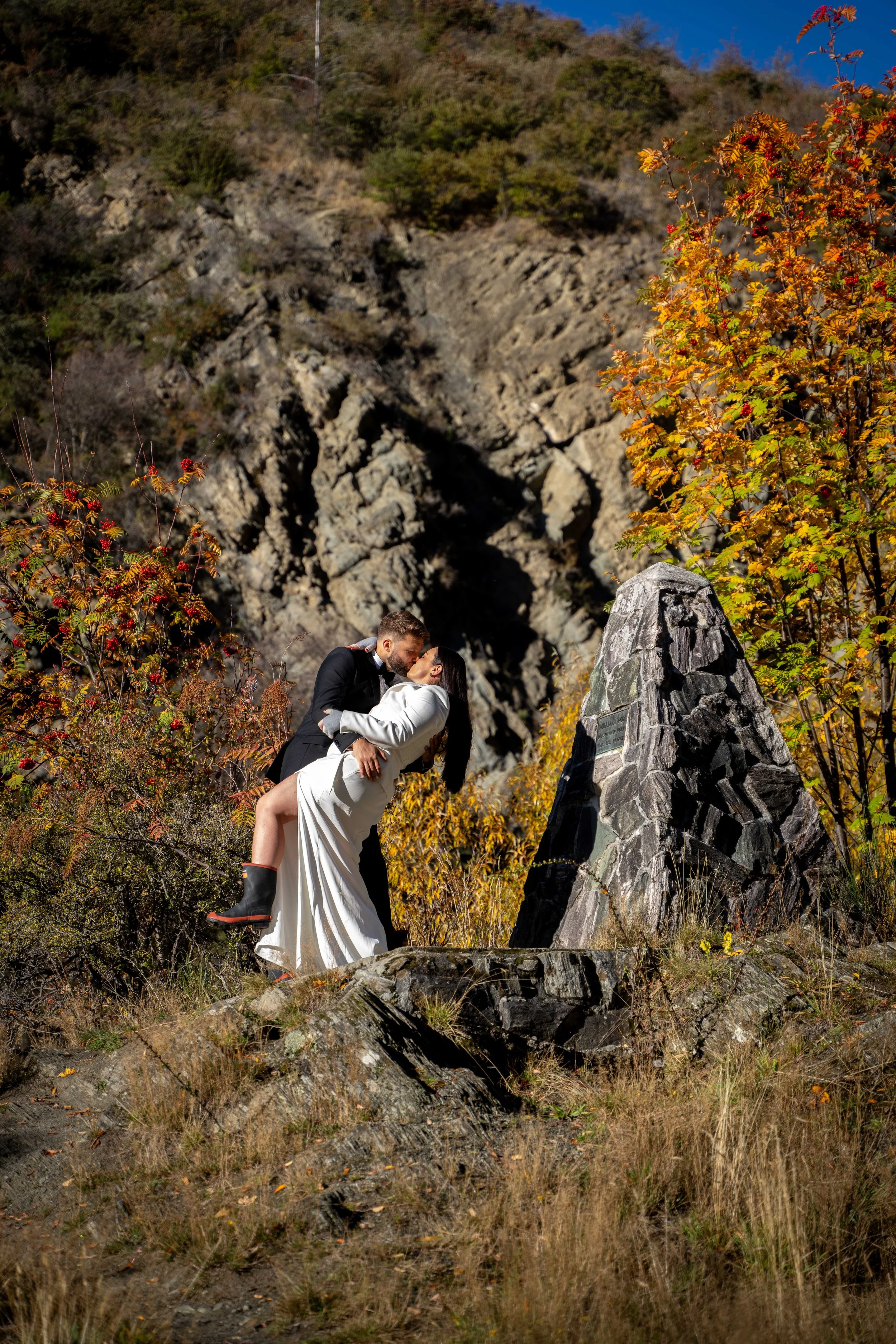 A couple, dressed formally with gumboots, sharing a kiss outdoors in front of a rocky monument with colourful autumn foliage in the background.
