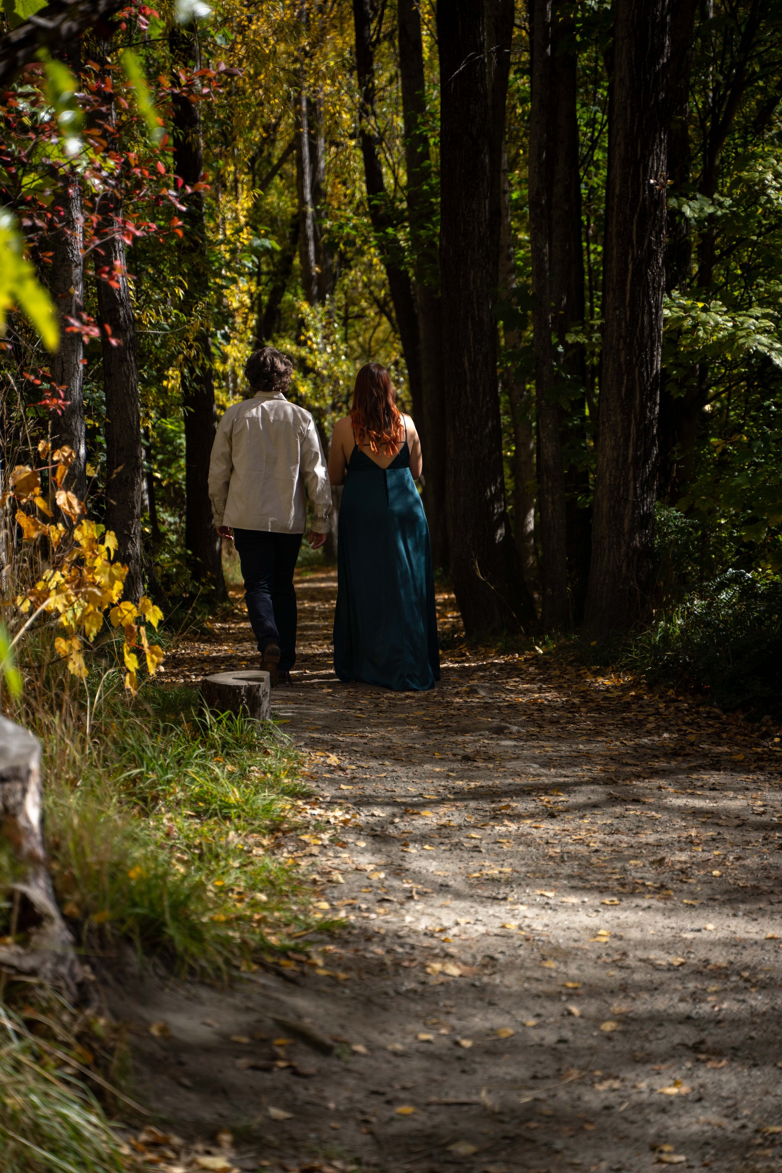 Two people walking along a forest trail during the daytime, surrounded by tall trees with sunlight filtering through the leaves.
