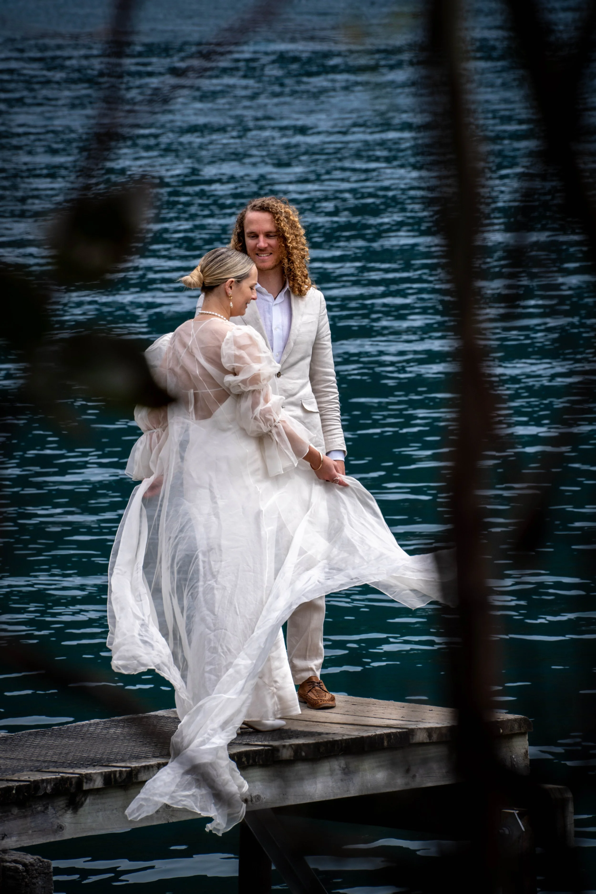 Couple in wedding attire standing on a wooden dock by a lake, celebrating their wedding.