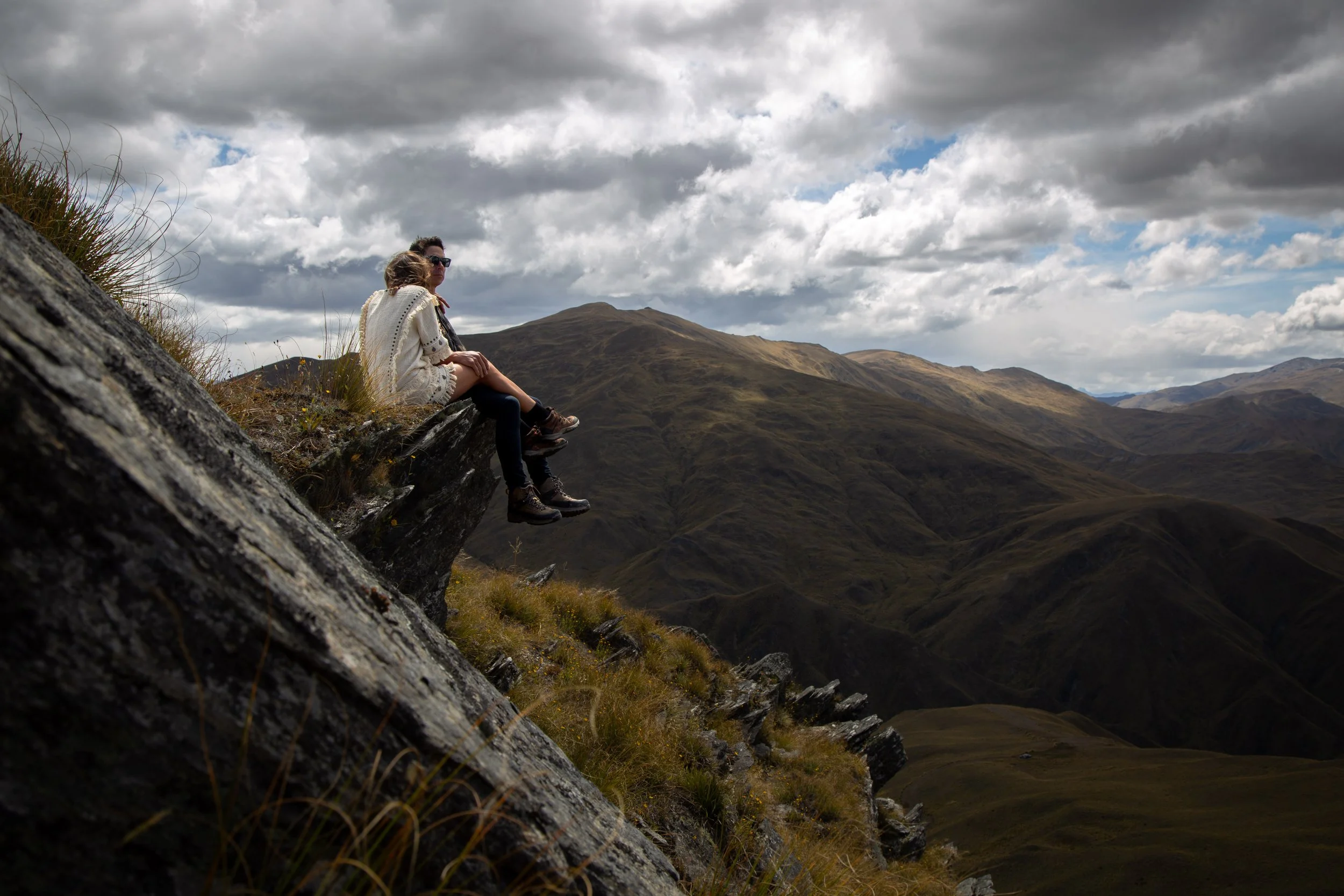 Two people sitting on a rocky ledge overlooking mountains and a cloudy sky.