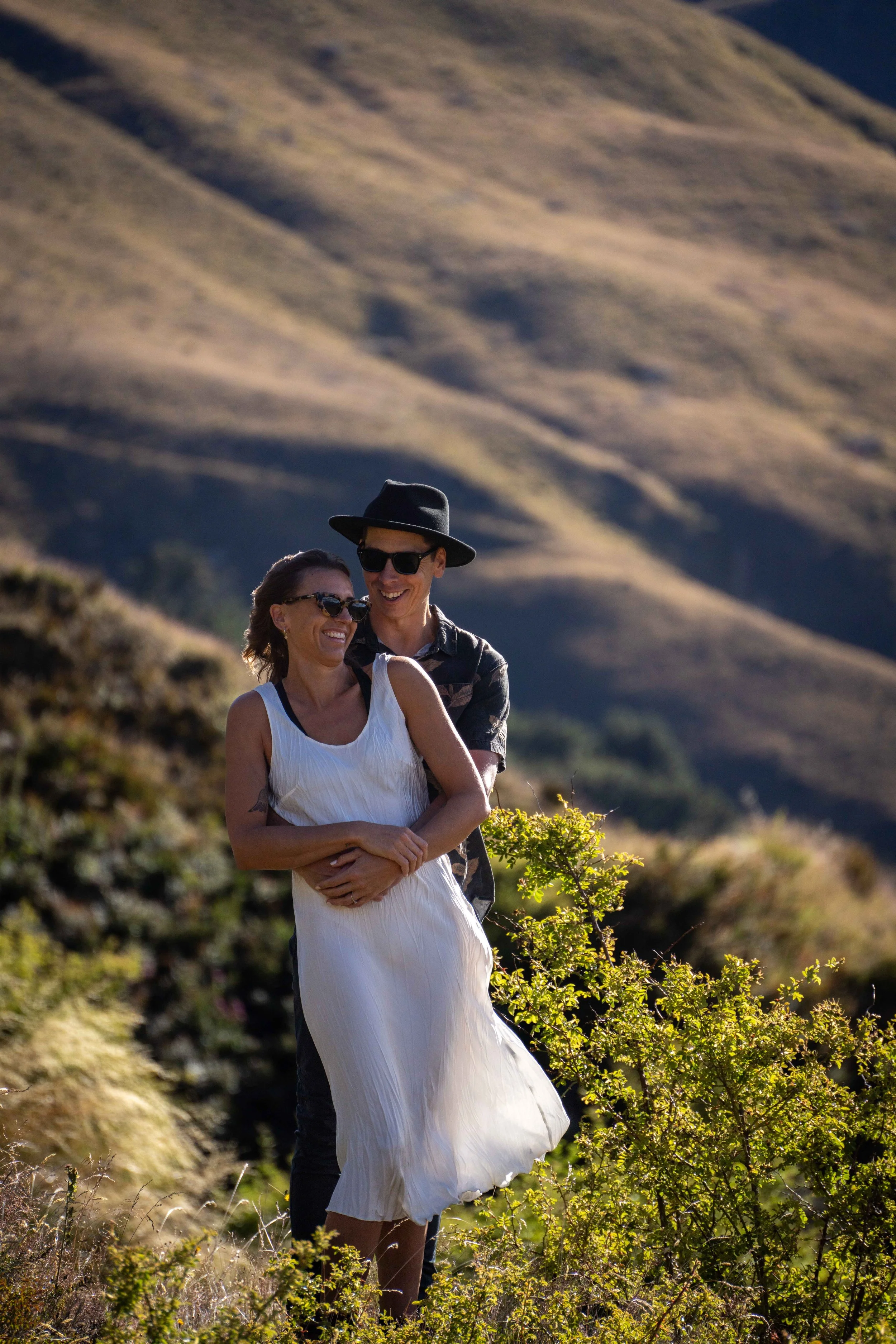 A smiling couple stands outdoors in a mountainous area with grassy hills in the background. The woman is wearing a white sleeveless dress and sunglasses, and the man is wearing a black hat, sunglasses, and a dark shirt, with the man hugging the woman