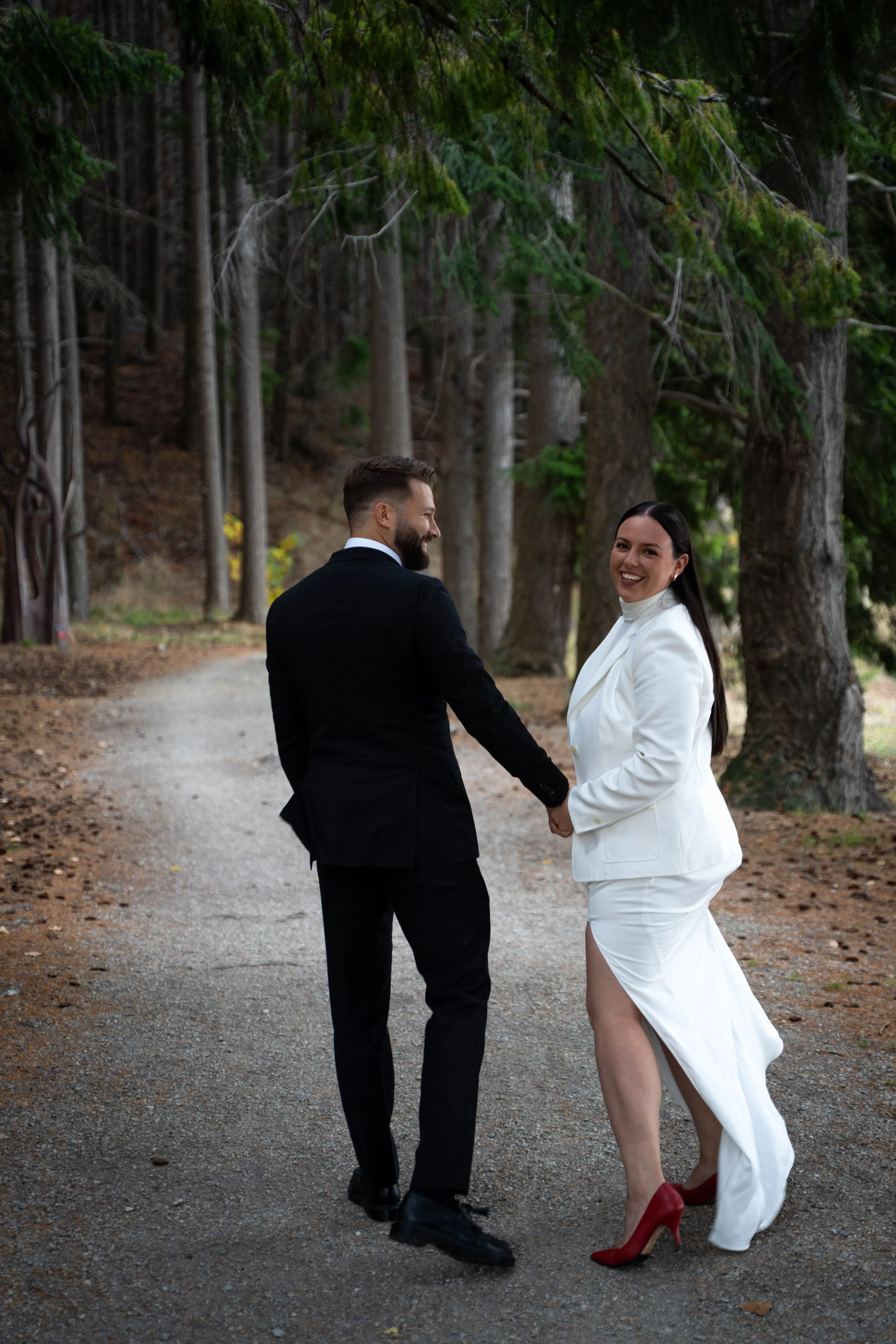 A couple dressed in wedding attire holding hands and walking through a forested trail, smiling at each other.