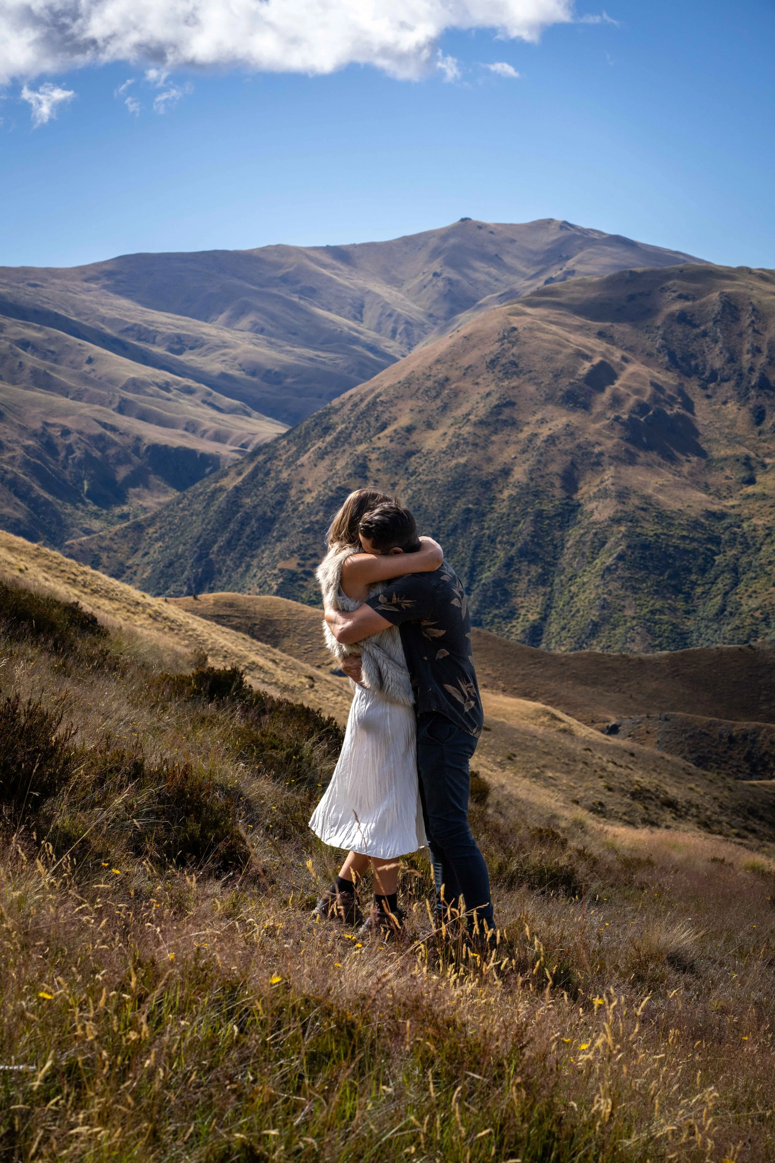 A couple hugging on a grassy hillside with mountains in the background during daytime.