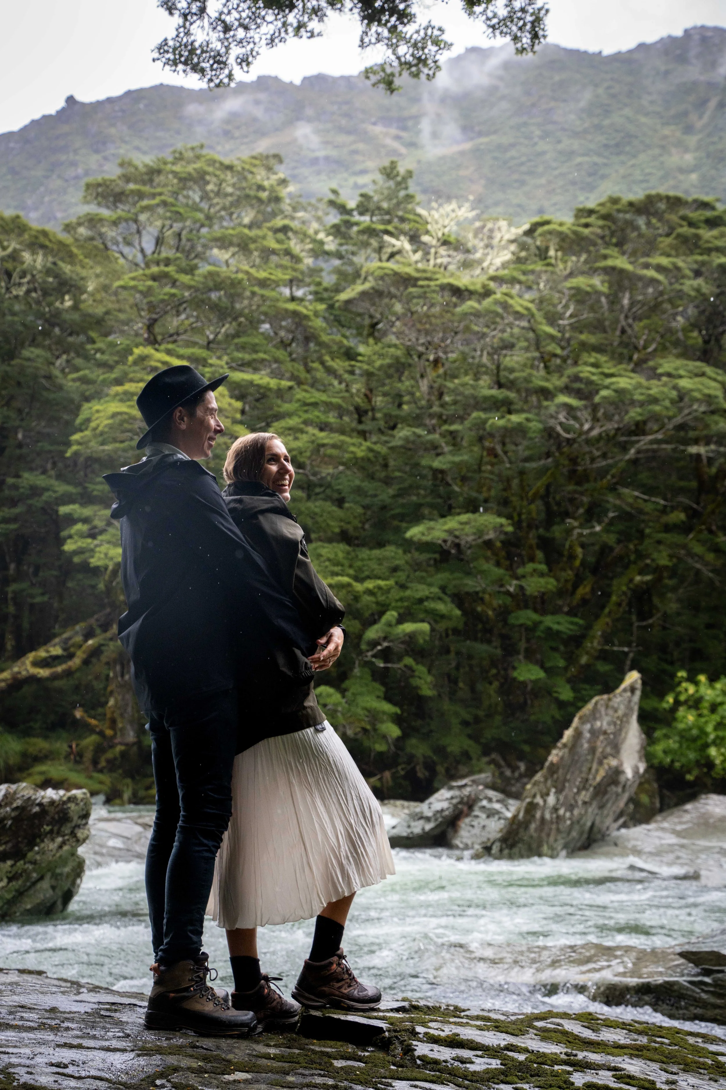 A man and woman standing on rocks by a river, smiling and looking into the distance, surrounded by lush green trees and mountains.