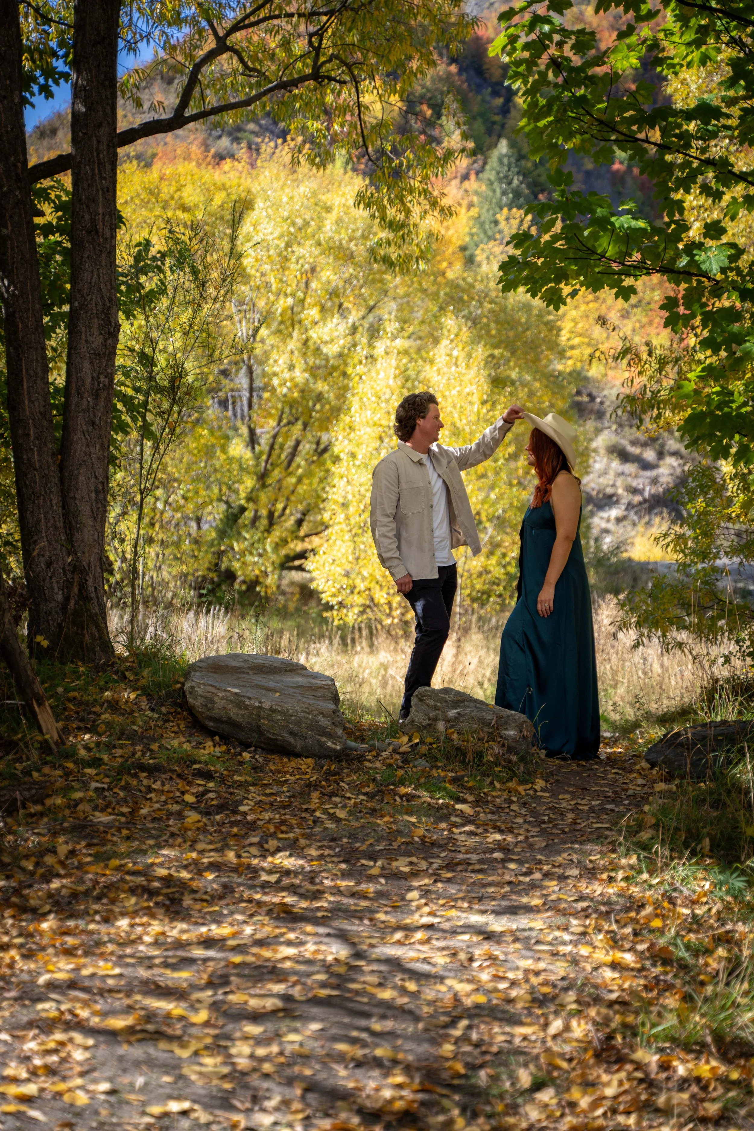 A couple standing in a forest with fall foliage, the man is touching the woman's hat as they look at each other.
