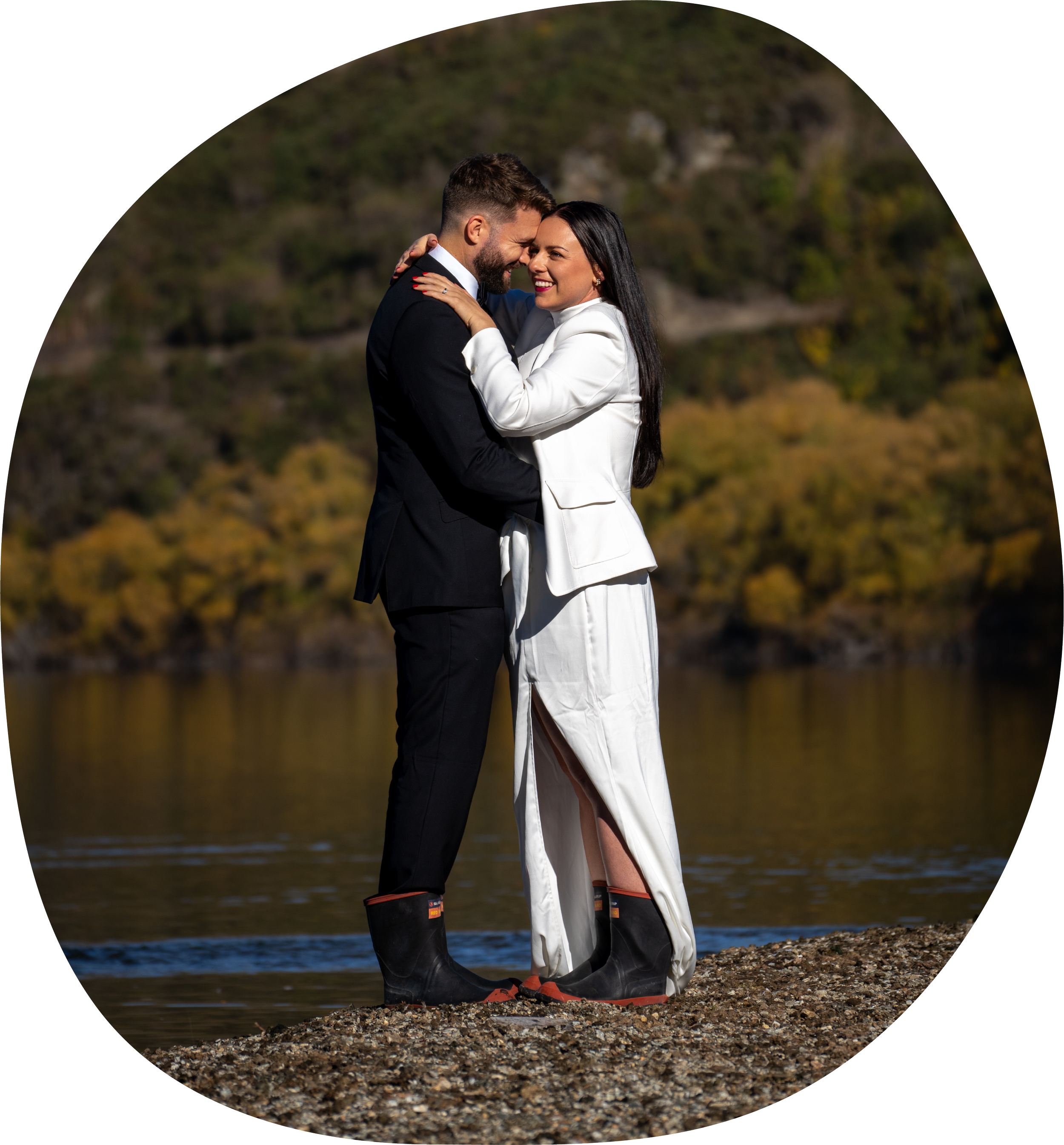 A couple dressed in wedding attire standing close together outdoors near water, smiling and embracing each other.