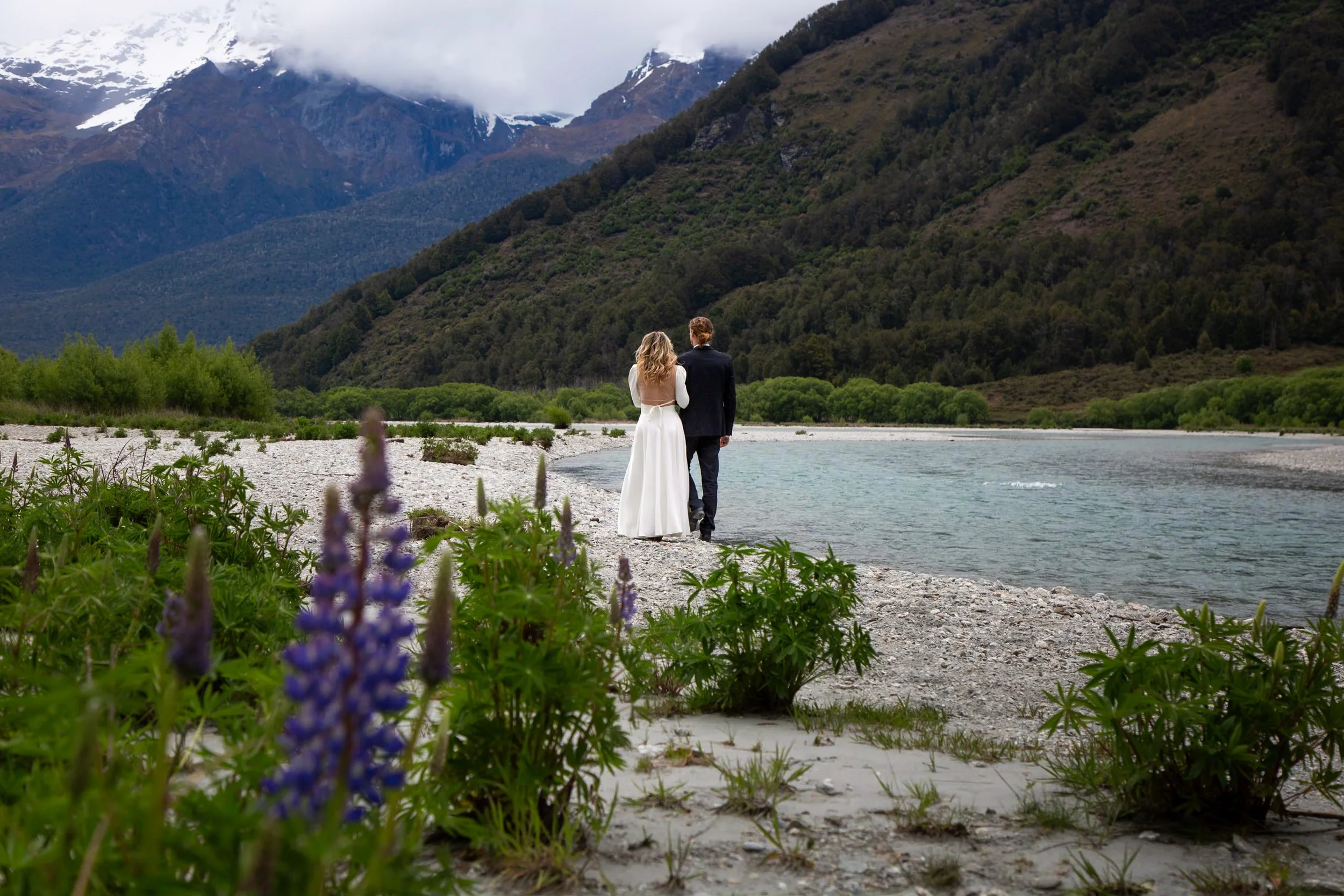 A couple, a bride in a white dress and a groom in a black suit, walk along a riverbank with mountainous landscape in the background, lush green trees, and cloudy sky.