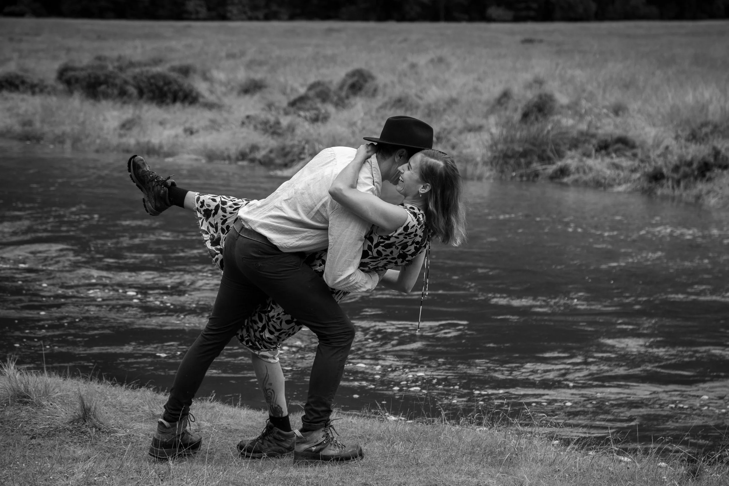 A man and woman by a river, with the man holding the woman as she leans back, both smiling and looking at each other, in a playful pose.