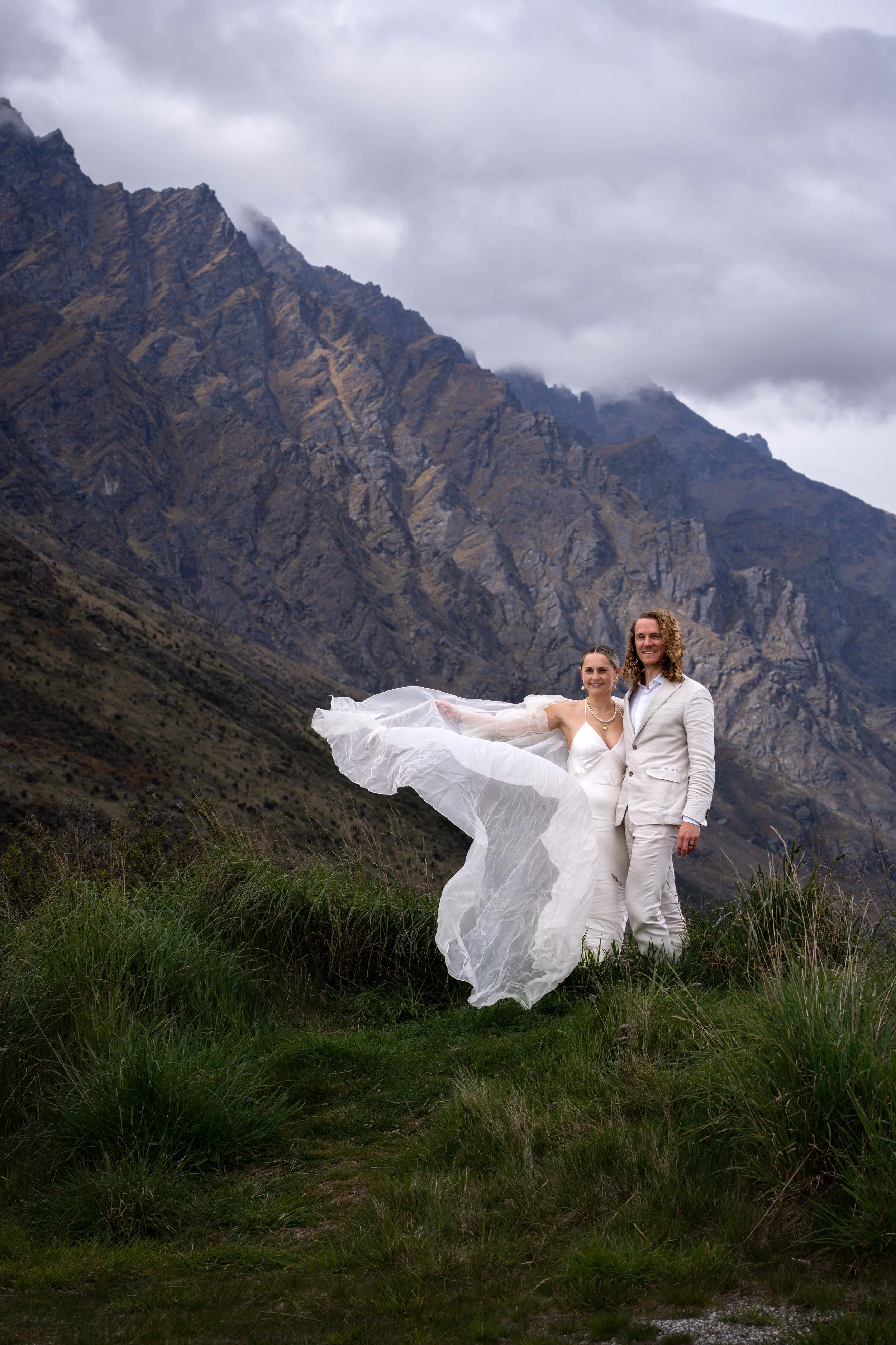 A bride and groom standing on grass in front of mountains, with the bride holding her flowing dress and the groom dressed in a white suit, on a cloudy day.