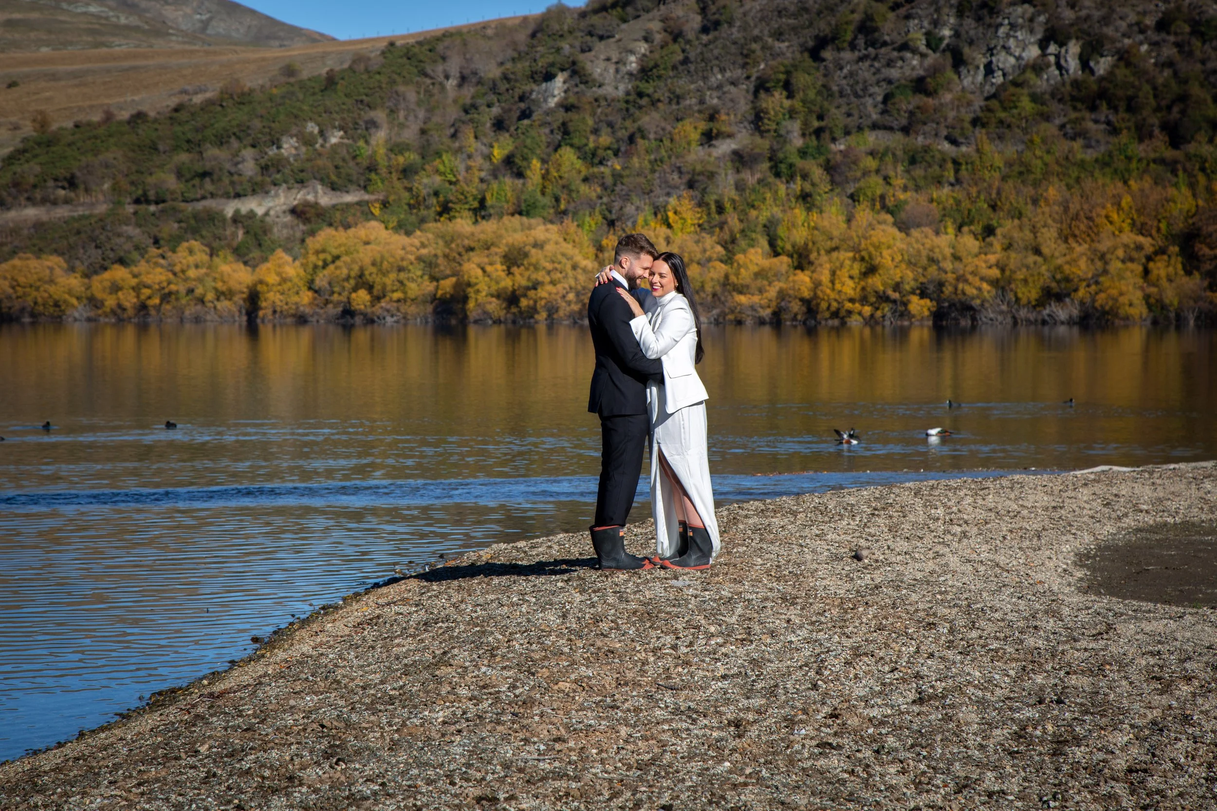 A couple dressed in wedding attire embraces on a rocky shoreline by a calm lake, with autumn-colored trees and hills in the background.