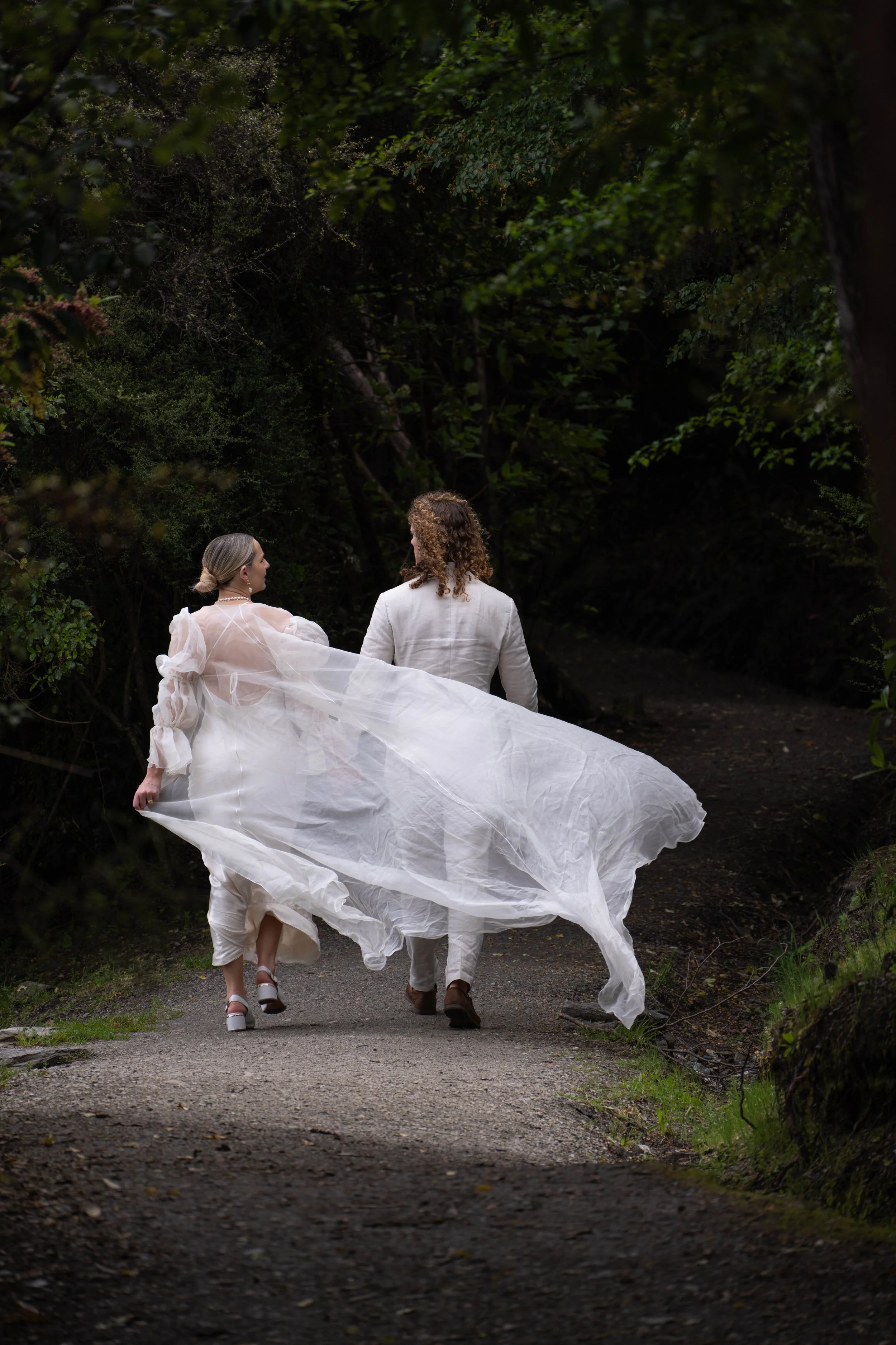 A couple walking on a forest trail, dressed in white wedding attire, with the bride's dress flowing.